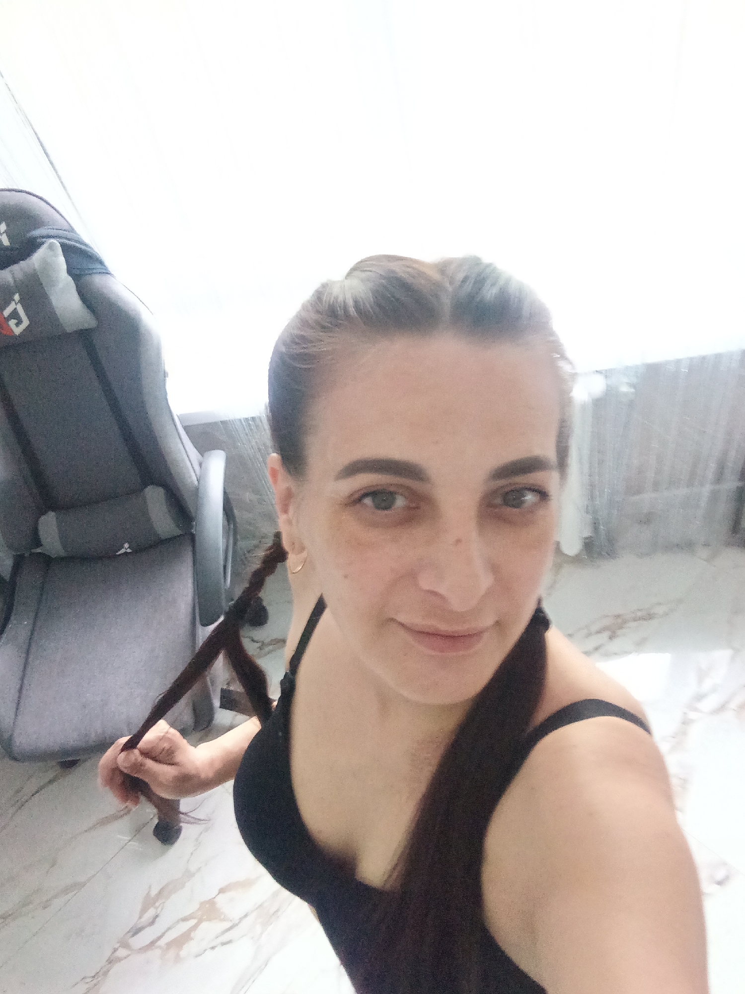 A woman is posing for a selfie indoors, next to a gray gaming chair. She is wearing a black tank top and has her hair in braids. The background includes a window covered with curtains. The floor is marble. The location is Unknown, Unknown.