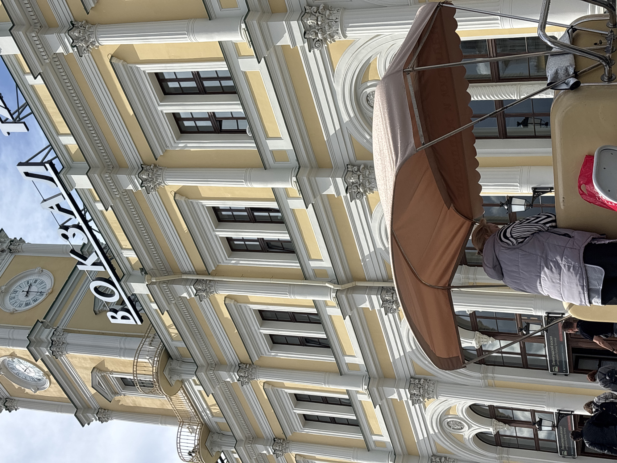 The image depicts the building of the House of Books ("Дом Книги") in Moscow, Russia. The building's facade is visible, showcasing its architectural details, including columns, ornate decorations, and windows. A clock tower with a large clock face is also visible. Part of a small tourist vehicle with an awning is in the foreground, and blurred people are present on the street. Signage referring to Leningradsky vokzal (Ленинградский вокзал, Leningrad Railway Terminal) is apparent in the vicinity. The environment suggests an urban setting, with a mix of architectural beauty and pedestrian activity.