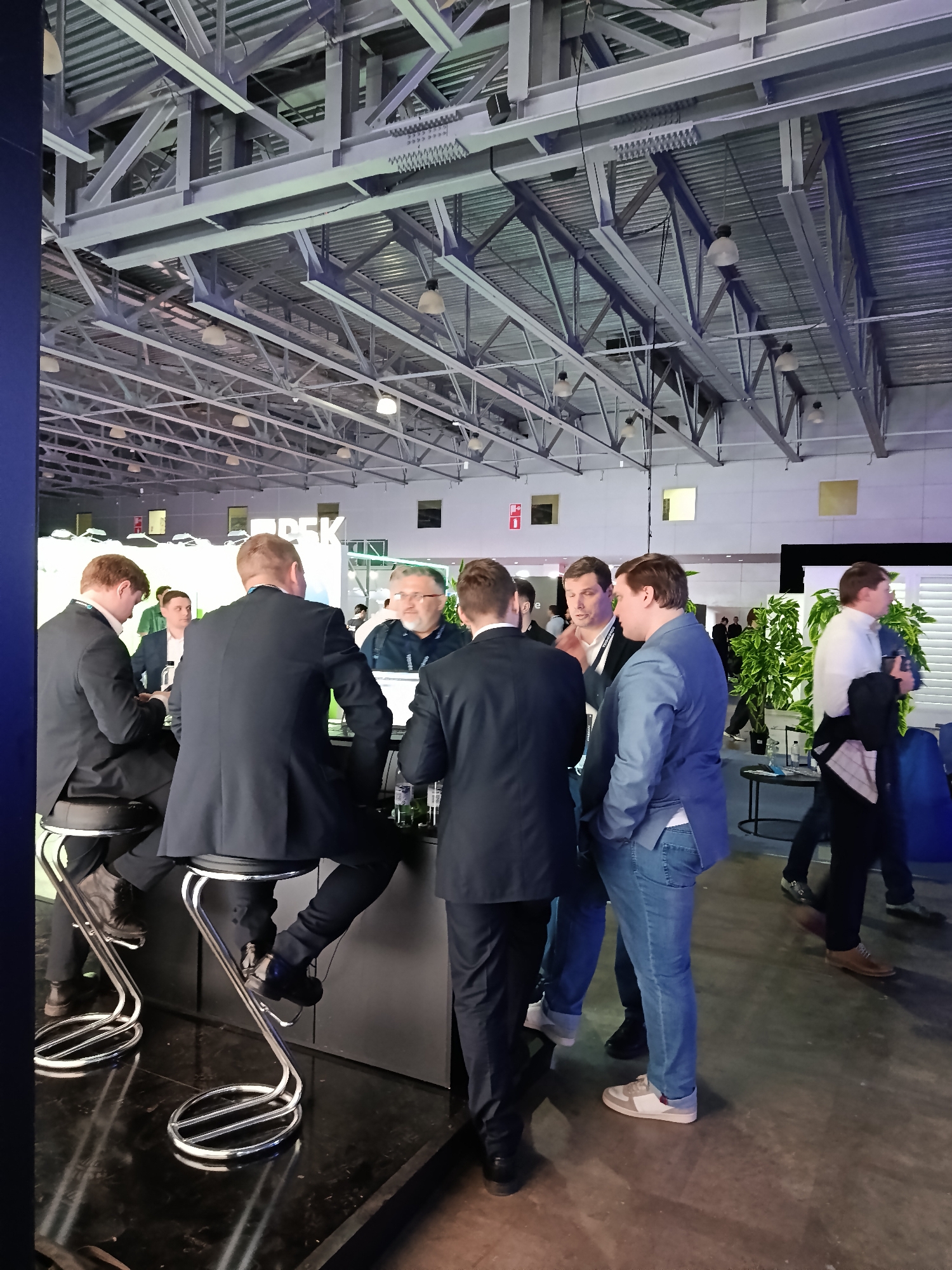The image shows an indoor event with several people gathered around a bar area. Six men are in focus, some wearing suits and others in more casual attire such as jeans and a blue jacket. Two men are seated on bar stools, while others are standing and interacting. A counter is visible, and behind the people are plants and other indistinct figures. The setting appears to be a convention or exhibition hall, evidenced by the high ceiling with exposed metal beams and industrial-style lighting fixtures. The presence of event booths and signage in the background suggests a business or promotional gathering. There is no definite information to indicate that the location is in the city of Khimki, Russia.