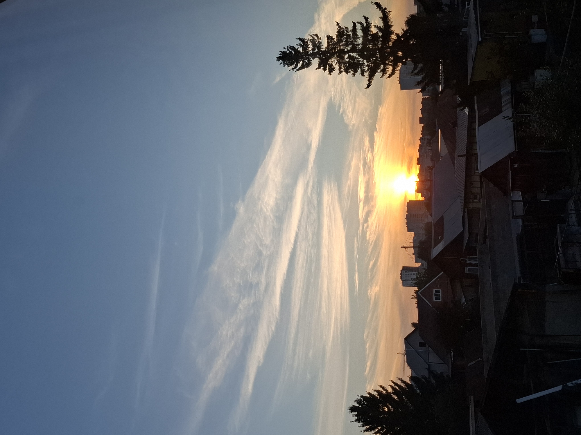The image depicts a sunset over a residential area in the city of Tyumen, Russia. The sun is a bright, glowing orb low on the horizon, casting a warm, orange light that reflects off the sky and illuminates the clouds. The sky is a gradient of deep blue at the top, transitioning to lighter blues and oranges closer to the horizon. Thin, wispy clouds stretch across the sky, adding texture and depth to the scene.
In the foreground, the silhouettes of various buildings are visible, including houses with pitched roofs and some taller structures in the distance. One prominent evergreen tree stands tall on the right side of the image, its dark silhouette contrasting with the bright sky. The lower part of the image shows darker, more shadowed structures, possibly outbuildings or sheds. The overall impression is a tranquil evening scene in a suburban or semi-urban environment.