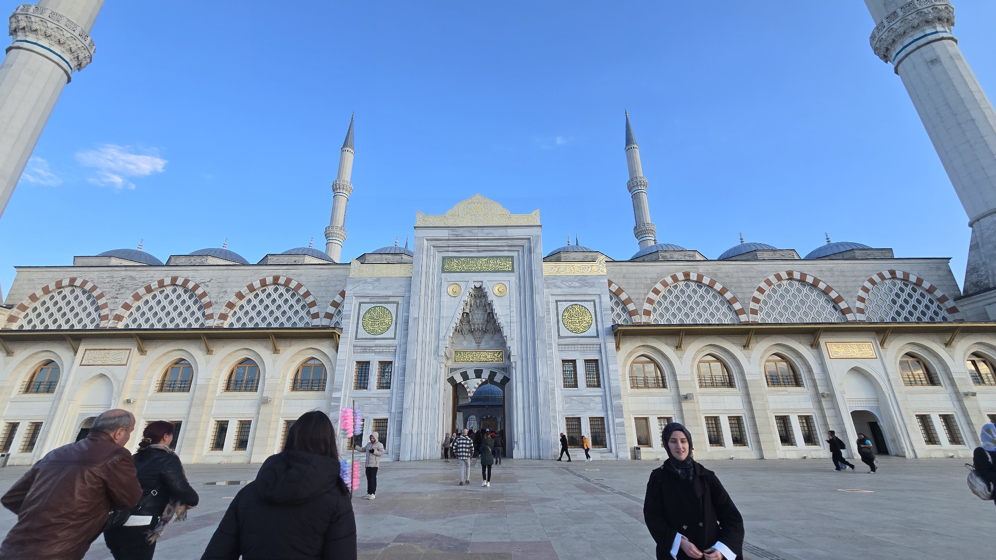 The image captures a sunny day in Üsküdar, Turkey, in front of the Çamlıca Mosque. The imposing structure of the mosque features minarets, domes, and a grand entrance adorned with Islamic calligraphy and patterns. Several people are present in the plaza. To the left, an older man and woman are walking. A woman in a black coat stands to the right of the frame. In the center, a few people are entering or exiting the mosque. The sky is clear and blue, providing a vibrant backdrop.