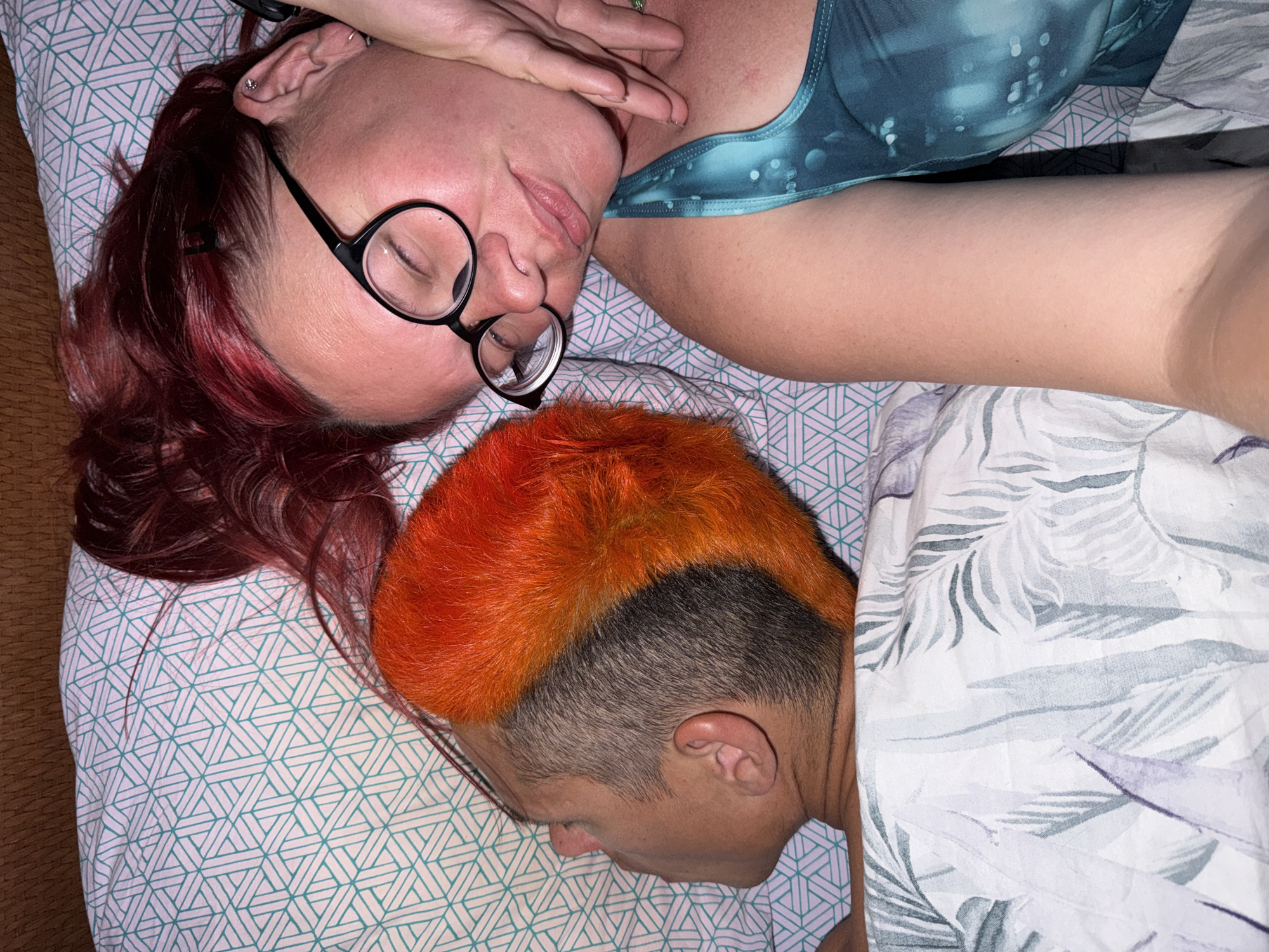Two individuals are lying on a bed covered with patterned sheets. One individual, possibly female, has red hair, wears glasses, and is positioned with their head near the top left of the frame. Their eyes are closed and one hand is near their face. They are wearing a green and blue patterned top. The second individual, possibly male, has orange hair styled in a Mohawk and is mostly covered by a floral patterned sheet. Their head is positioned near the bottom of the frame. The location is unspecified and cannot be determined from the image. The city of Chaikovsky, Russia is not evident in this image. The individuals appear to be sleeping.