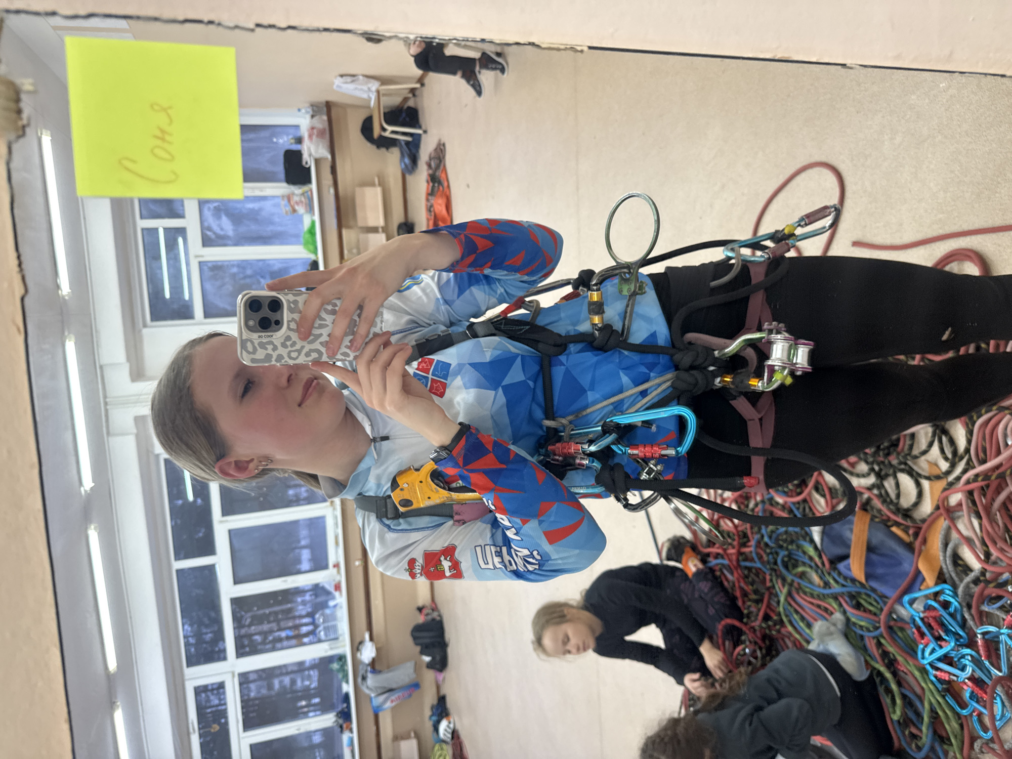 A young woman takes a selfie in front of a mirror. She is wearing climbing gear, including a harness and carabiners. In the mirror, a yellow sticky note with the name "Соня" (Sonya) written in Russian is visible. The reflection also shows a window with multiple panes and other objects in the background. To the right of the woman, climbing ropes and other people are visible. The image suggests the location is Berezniki, Russia, due to the Cyrillic lettering on the woman's sleeve.