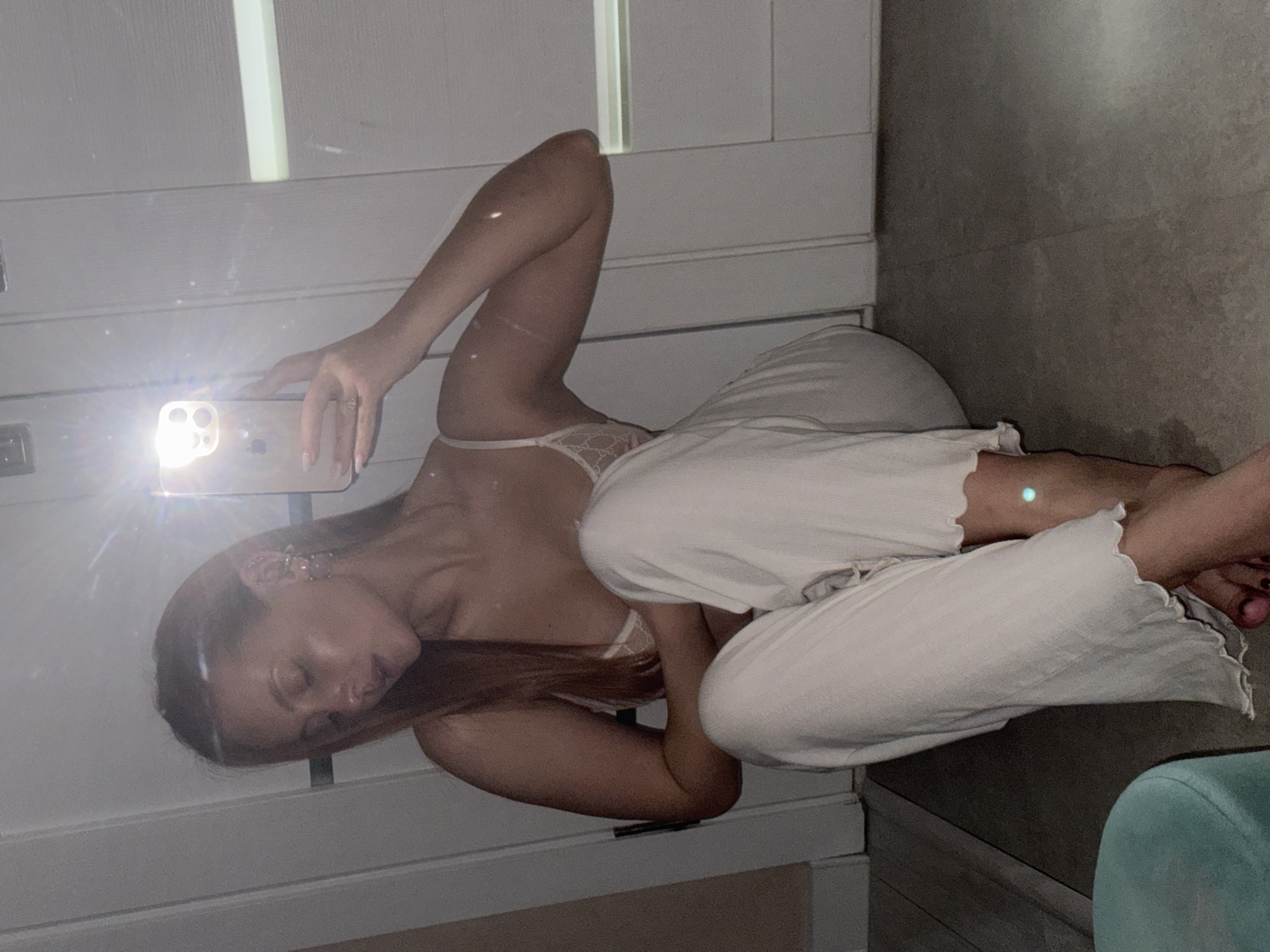 A woman is taking a selfie in a mirror. She is wearing white pants and a light-colored top. The background consists of a white wall, a grey tiled floor, and a blue chair. It is not possible to determine the location in the city of Chaikovsky, Russia.