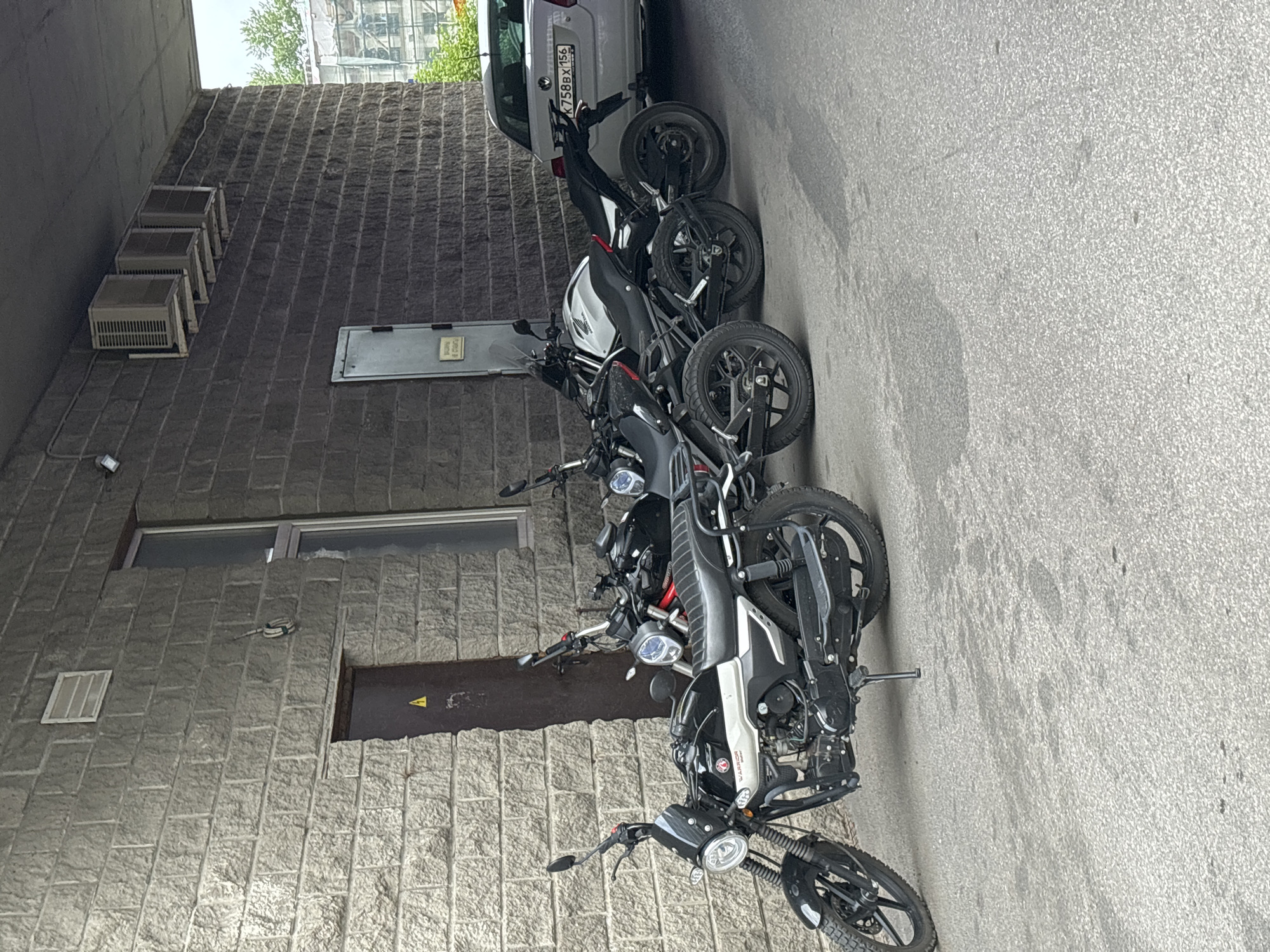 The image shows a row of parked motorcycles next to a brick building in an urban environment. The building features a window, a door with a caution symbol, and several air conditioning units. A silver car is parked adjacent to the motorcycles. The street surface is paved with asphalt. The license plate of the car reads "K758BX156".