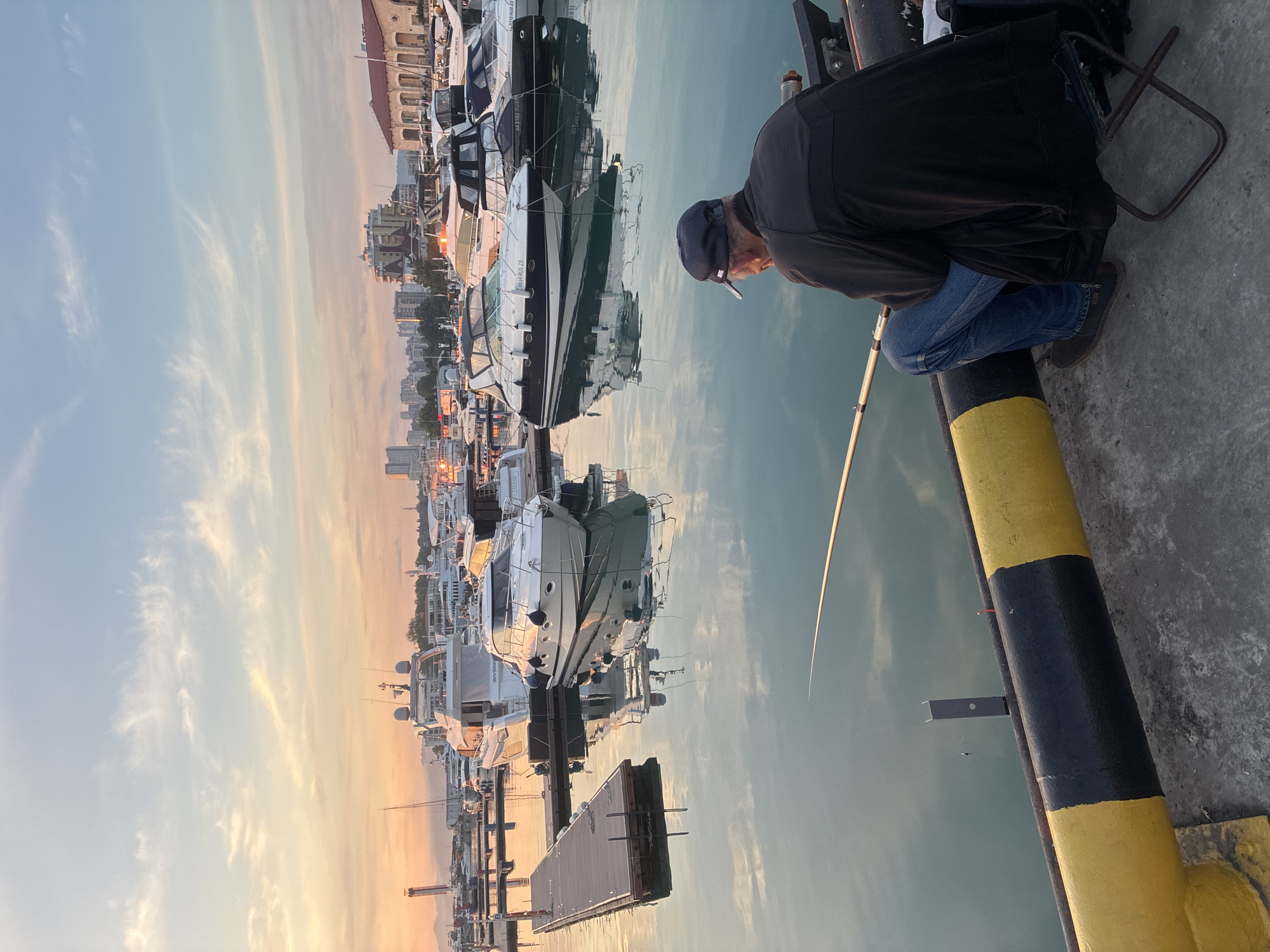 The image depicts a marina at what appears to be either sunrise or sunset in the city of Sochi, Russia. A man, positioned with his back to the viewer, is seated on a small metal folding stool at the edge of a concrete dock. He wears a dark jacket, blue jeans, and a dark baseball cap with a white "Nike" logo. He holds a fishing rod, extending its tip into the calm, reflective water of the marina.
The dock's edge in the immediate foreground features a thick, cylindrical bumper painted with alternating black and yellow stripes. To the left, a wooden floating walkway with metal posts extends into the water.
The marina is filled with numerous docked boats, primarily white motor yachts and cruisers of varying sizes. Reflections of the boats and the sky are visible on the water's surface. Several boats display Russian registration numbers, such as "0634 RUS 23" and "AA3102 RUS 23," with "RUS 23" corresponding to Krasnodar Krai, where Sochi is located.
In the background, a city skyline stretches along the coastline. It includes multi-story buildings, some modern high-rises, and older architectural structures, including a prominent building with a red-tiled roof. Distant mountains are visible on the horizon to the left, and a large crane structure is also present in that direction.
The sky exhibits a gradient of color, transitioning from a warm orange and yellow near the horizon to a pale blue overhead, with scattered, wispy white clouds. The overall lighting suggests the golden hour, casting soft, warm tones across the scene.