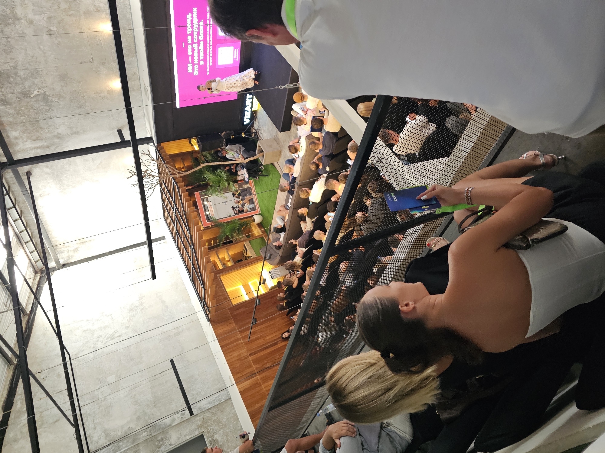 Captured from an elevated perspective, the photograph reveals a presentation or conference in progress at an indoor venue. The location is identified as North Kuta, Indonesia. A diverse group of people, dressed in casual and business attire, occupy the audience, with some seated and others standing. A presenter is visible on stage, speaking into a microphone. The backdrop features a large screen displaying the VIZART logo and a text banner in a non-English language. Attendees are capturing the event using their mobile devices, while others engage in conversation. The setting incorporates wooden architectural elements, plants, and a modern design aesthetic. A woman, situated in the foreground and closer to the viewer, is holding a pamphlet or document. The photograph provides an overview of a professional event taking place in a contemporary setting in North Kuta.