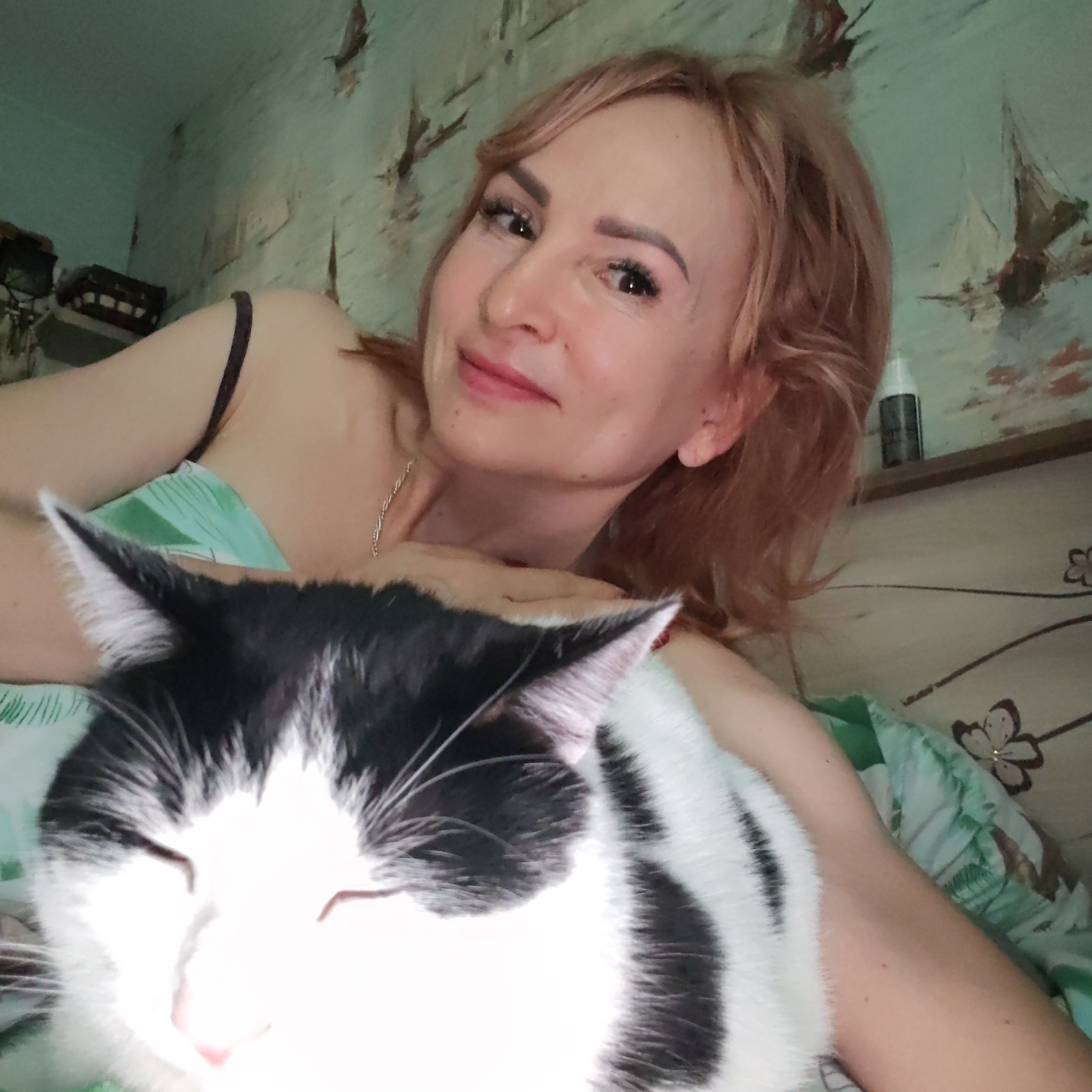 A woman with auburn hair is lying in bed, looking at the camera with a slight smile. She is wearing a black spaghetti strap top and a silver necklace. A black and white cat is lying on her chest. The bed has green floral patterned sheets. Behind the woman is a shelf with a small black bottle, and a wall with a pattern of sailboats. The location is Chaikovsky, Russia.