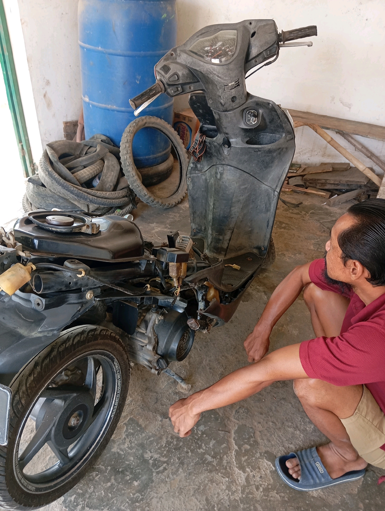 A man in a red polo shirt and khaki shorts kneels on a concrete floor, looking at a disassembled black scooter. He has dark hair and a goatee. The scooter is missing body panels, exposing its engine and wiring. In the background, a large blue barrel sits next to a pile of old tires and wooden planks. The scene appears to be an outdoor workshop or storage area.