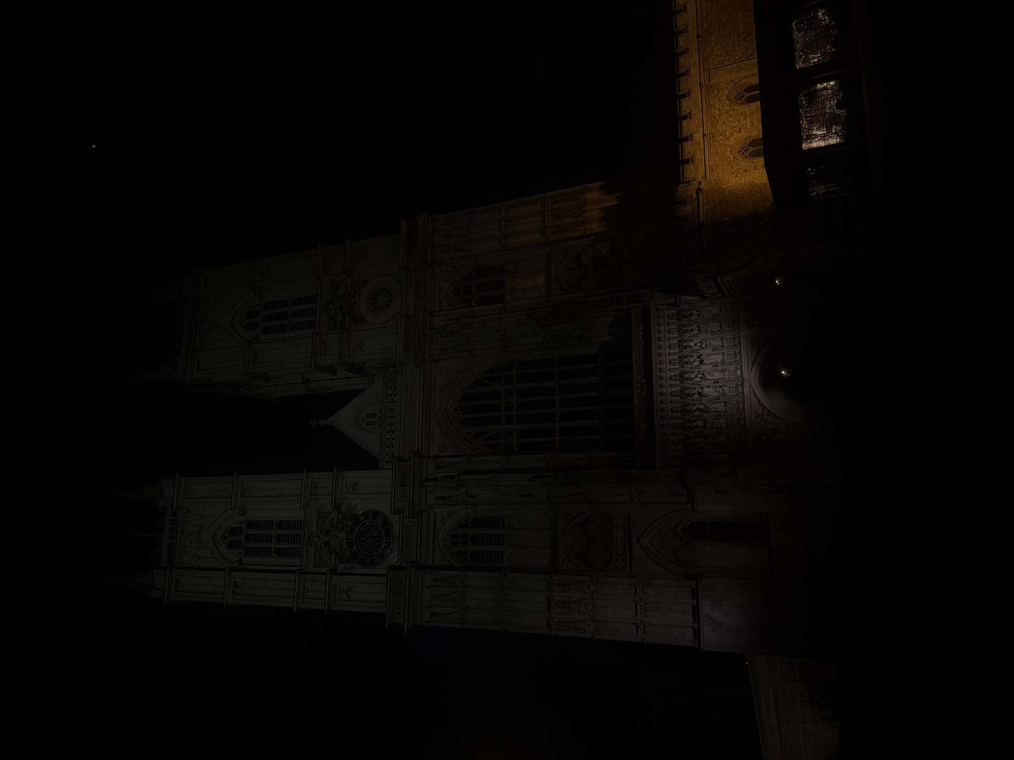 The image depicts Westminster Abbey at night in London, United Kingdom. The abbey is illuminated, showcasing its Gothic architecture, including pointed arches, large windows, and intricate stonework. The scene is dark, with the Abbey appearing as a lighter, almost ghostly structure against the black sky. A tall tower is visible on the right side. The focus is on the building's facade, emphasizing its architectural details.
