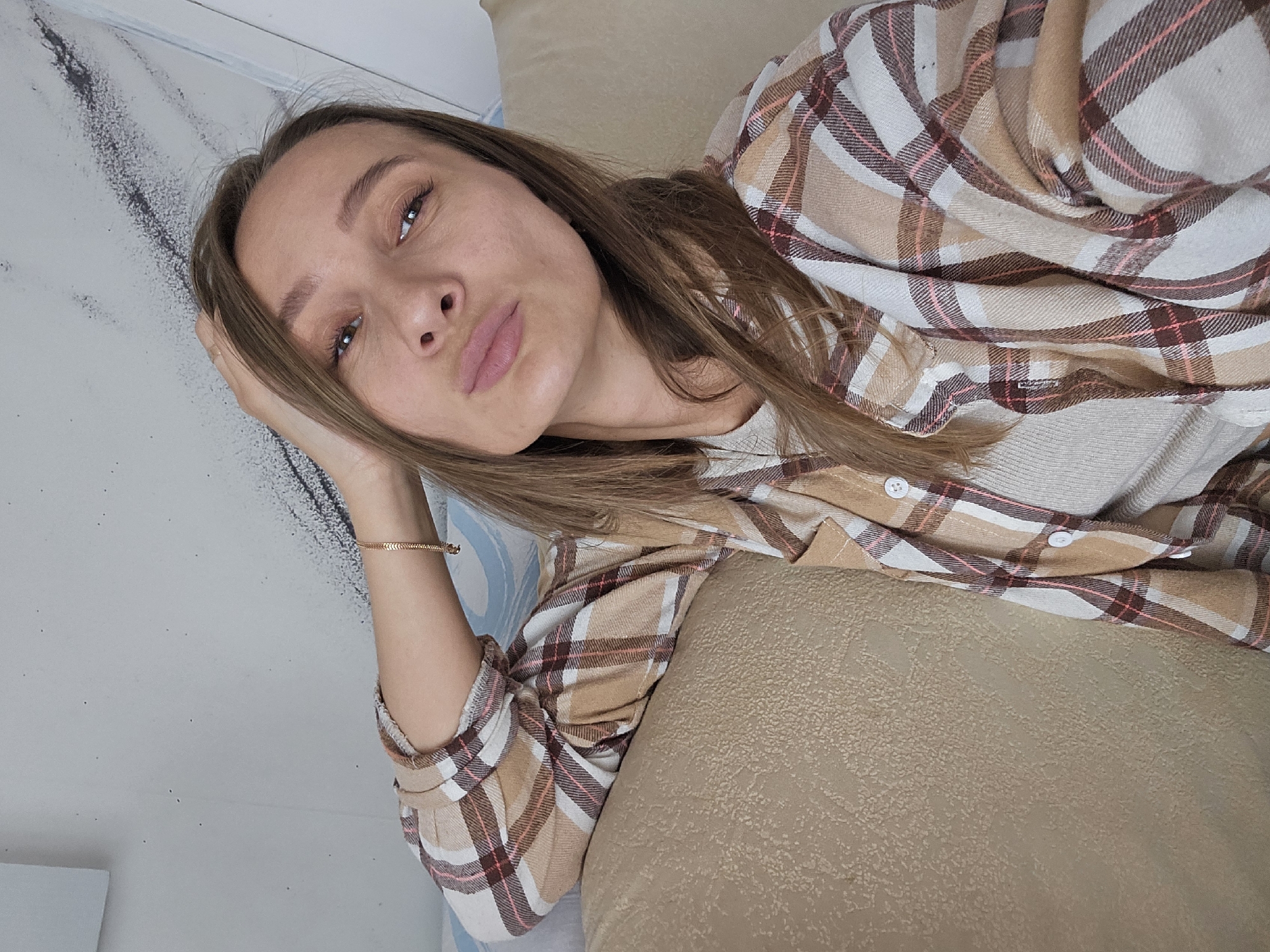 An indoor selfie captures a woman reclining on a light brown, textured surface, likely a sofa, in the city of Chaikovsky, Russia. She gazes directly into the camera with a pouting expression, her light brown hair falling over her shoulders. Her left hand is positioned near her head, showcasing a thin gold-colored chain bracelet on her wrist. She wears an open plaid flannel shirt featuring a pattern of tan, white, dark brown, and thin pink stripes, layered over a light beige ribbed top. The background is a light gray wall adorned with a prominent, dark, abstract streaky pattern resembling paint strokes or marble, interspersed with small dark speckles. A hint of a light blue pattern is visible to her upper left, and a white vertical element appears on the far right.