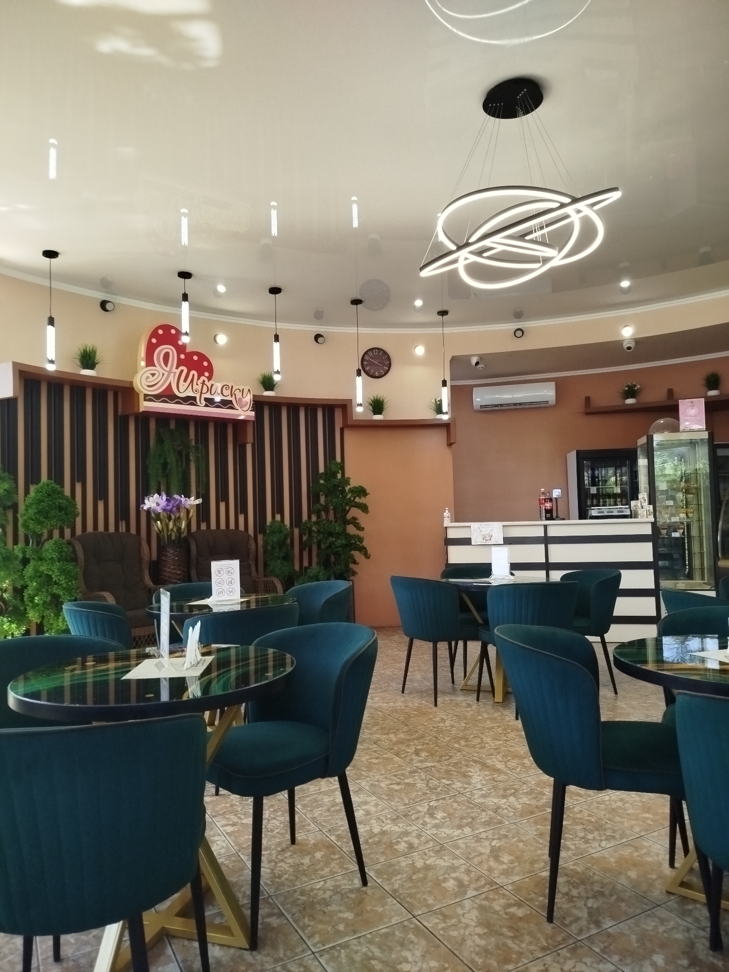 This is an interior photograph of a cafe in Taganrog, Russia. The cafe features a modern and elegant design with a light-colored, curved ceiling and recessed lighting. In the foreground, several dark teal upholstered chairs with black legs surround round, dark-topped tables with gold crisscross bases. The tabletops appear to have a glossy, reflective surface with a green and black swirling pattern. In the middle ground, there is a seating area with two plush, dark brown armchairs. Behind these chairs is a wall with vertical wooden slats and a decorative sign with two red hearts and the word "Ярмску" in stylized script, illuminated from behind by a warm light. Green artificial plants are strategically placed around this area.
Further back, a counter area is visible with a white and dark striped facade. On the counter, there are several bottles, a hand sanitizer dispenser, and some white display stands. Behind the counter, there is a glass-fronted refrigerator displaying drinks and a larger display case with glass shelves, likely for pastries or other food items. A white air conditioning unit is mounted high on the brown wall above the counter. A large, contemporary chandelier with multiple interconnected rings of light hangs from the ceiling, drawing attention to the central space. A round clock with a Roman numeral face is visible on the wall to the right of the decorative sign. The floor is tiled with a light brown, speckled pattern. There are no people visible in the image. The overall impression is of a clean, well-lit, and contemporary cafe.
