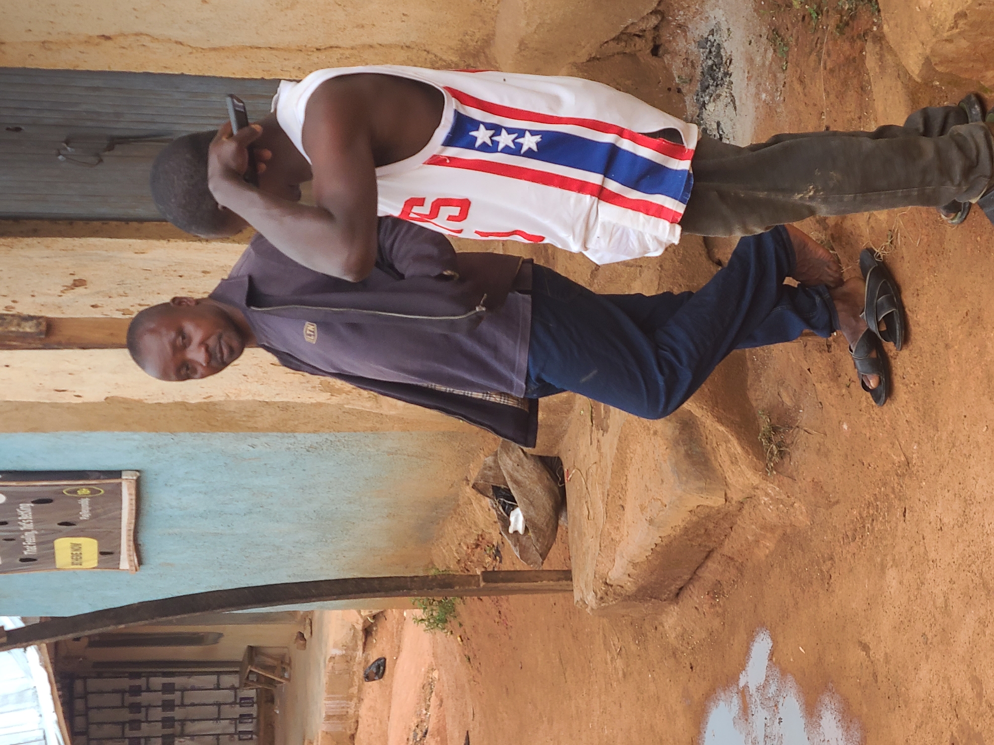 Two men are standing outdoors on a reddish-brown dirt ground, possibly in a residential or semi-urban area in Jos, Nigeria. The image is captured in daylight, under what appears to be overcast conditions or with diffused light.
In the foreground, a man with a dark complexion, wearing a dark blue zip-up jacket and blue trousers, looks directly towards the camera with a neutral expression. He is barefoot, but black sandals are visible on the ground near his feet.
Slightly behind him and to his right, another man with a dark complexion stands. He is wearing a white sleeveless tank top adorned with a distinctive design featuring red and blue horizontal stripes and three white stars on the blue stripe. The number "S5" is also visible in red on the white portion of his shirt. This man is holding a dark-colored cell phone to his right ear, seemingly engaged in a call, and is wearing dark sandals. He is wearing dark trousers.
The background features a light blue painted wall on the left, with a black power strip or charging station mounted on it. Visible text on this object includes "That Feeling, He's Reckless" (partially obscured) and "FREE NOW," along with a partial logo "Paigoo...". Above this blue wall is a yellowish-brown wall. To the right, a dark, possibly metal, door is visible, and further to the right, what appears to be a woven basket is on the ground. The ground itself is uneven, with patches of light-colored paint or residue and small bits of debris.