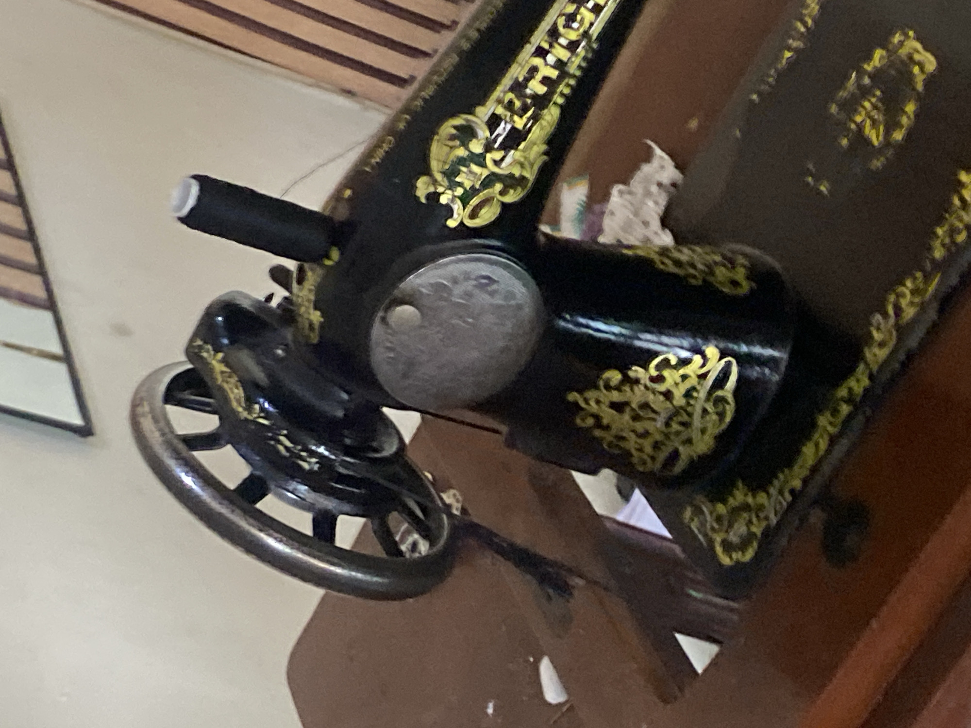 An antique black sewing machine with gold ornate decorations sits on a dark wooden table. The sewing machine features a large metal handwheel on the left and a black spool of thread on a spindle towards the back. The machine is positioned on a dark wooden table, with a faint white cloth or paper visible beneath it. The background consists of a light-colored wall and vertical wooden blinds. The scene is indoors, likely in a residential setting. The image does not contain any people or specific geographical landmarks to indicate its location within Jauro Yinu, Nigeria.