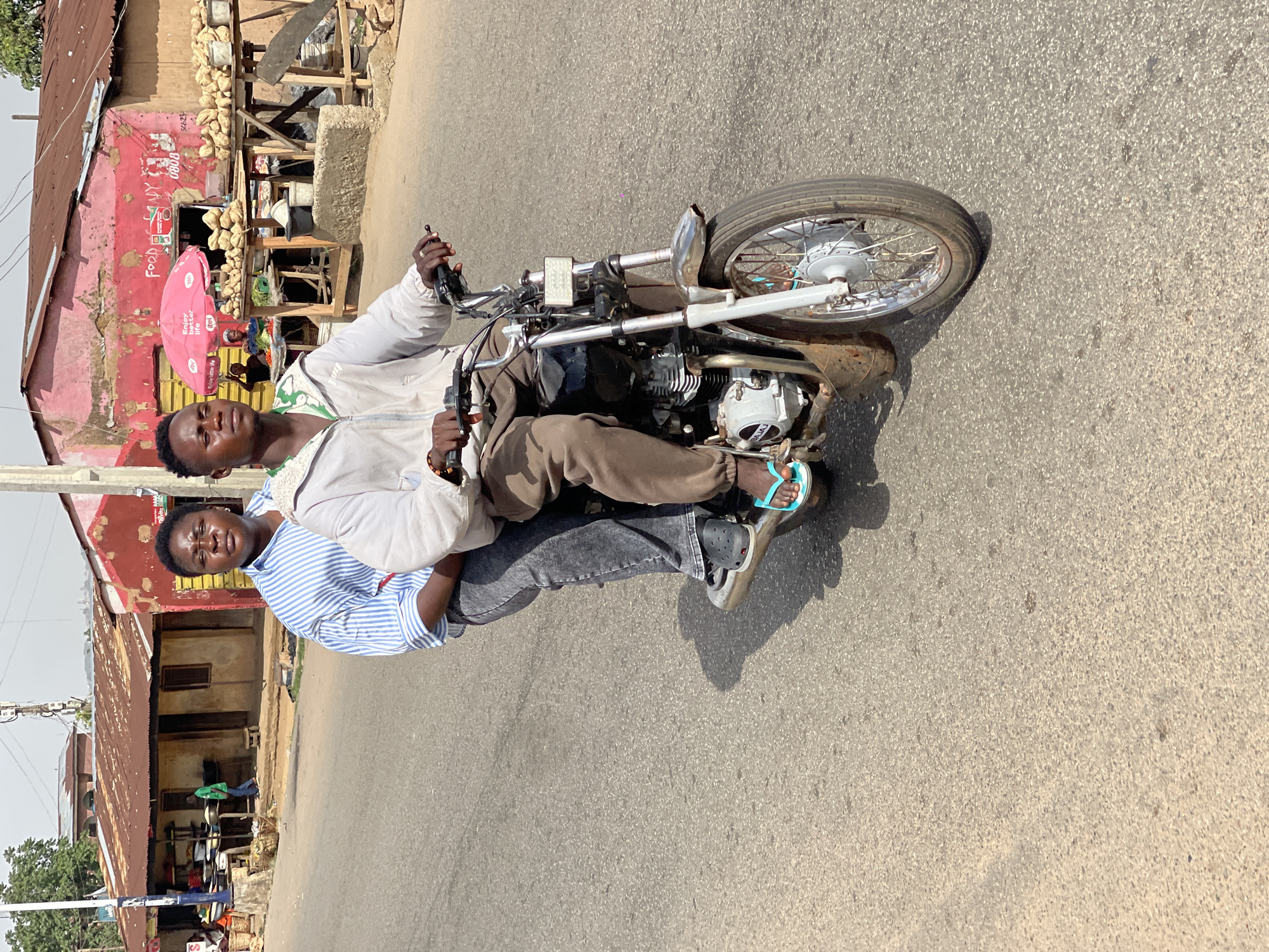 Two young men ride a motorcycle on a paved road in Jos, Nigeria. The driver is fair-skinned and wears a white jacket with a green collar. The passenger behind him is dark-skinned and wears a blue-striped shirt and gray pants. Both wear sandals. The background shows a weathered building with a red and pink facade with a red umbrella. The building appears to house a food stall.