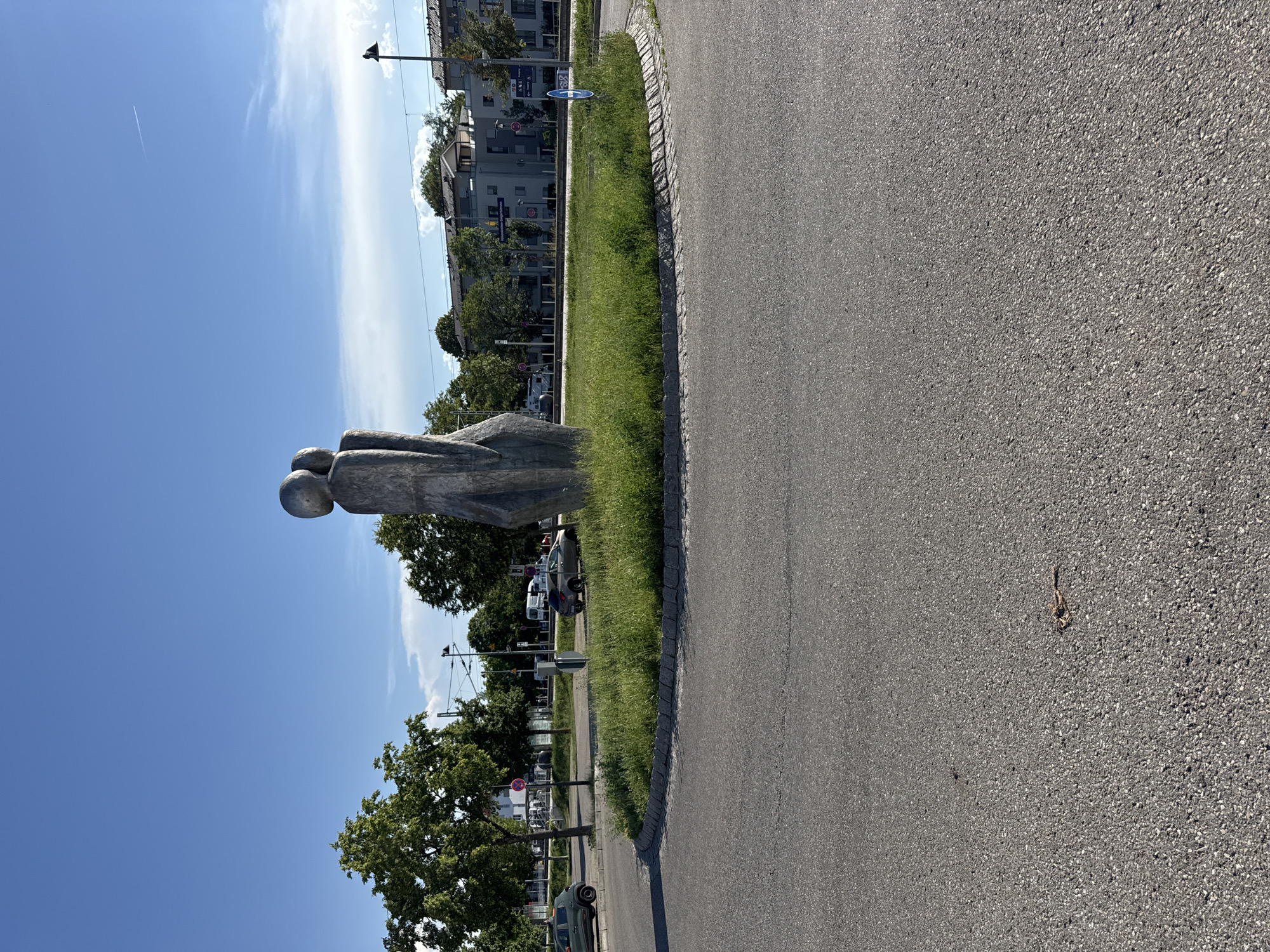 This is a landscape photograph, taken outdoors in a sunlit environment. The image is oriented vertically. In the foreground, a grey asphalt road curves to the right. A strip of green grass borders the road, separated by a cobblestone edge.
On the grass, a large, abstract sculpture is visible. It appears to be made of a dark, textured material and depicts two figures in a close embrace, with rounded heads.
In the background, there are buildings with multiple stories, some with balconies. Trees with green foliage are scattered throughout the scene. The sky is a clear, bright blue with a few wispy white clouds. A contrail from an airplane is faintly visible in the sky.
The scene suggests a public space, possibly a park or a street with artistic installations, in a suburban or urban setting. The bright sunlight and clear sky indicate it is daytime, likely midday or early afternoon. There are no discernible emotions conveyed by the sculpture or the overall scene, which appears peaceful and ordinary.
Visible text includes street signs with German text and arrows, as well as a blue circular sign with a bicycle icon, indicating a bike path or lane. There are also red and white circular signs that appear to be parking restrictions. The location is identified as Höhenkirchen-Siegertsbrunn, Germany.