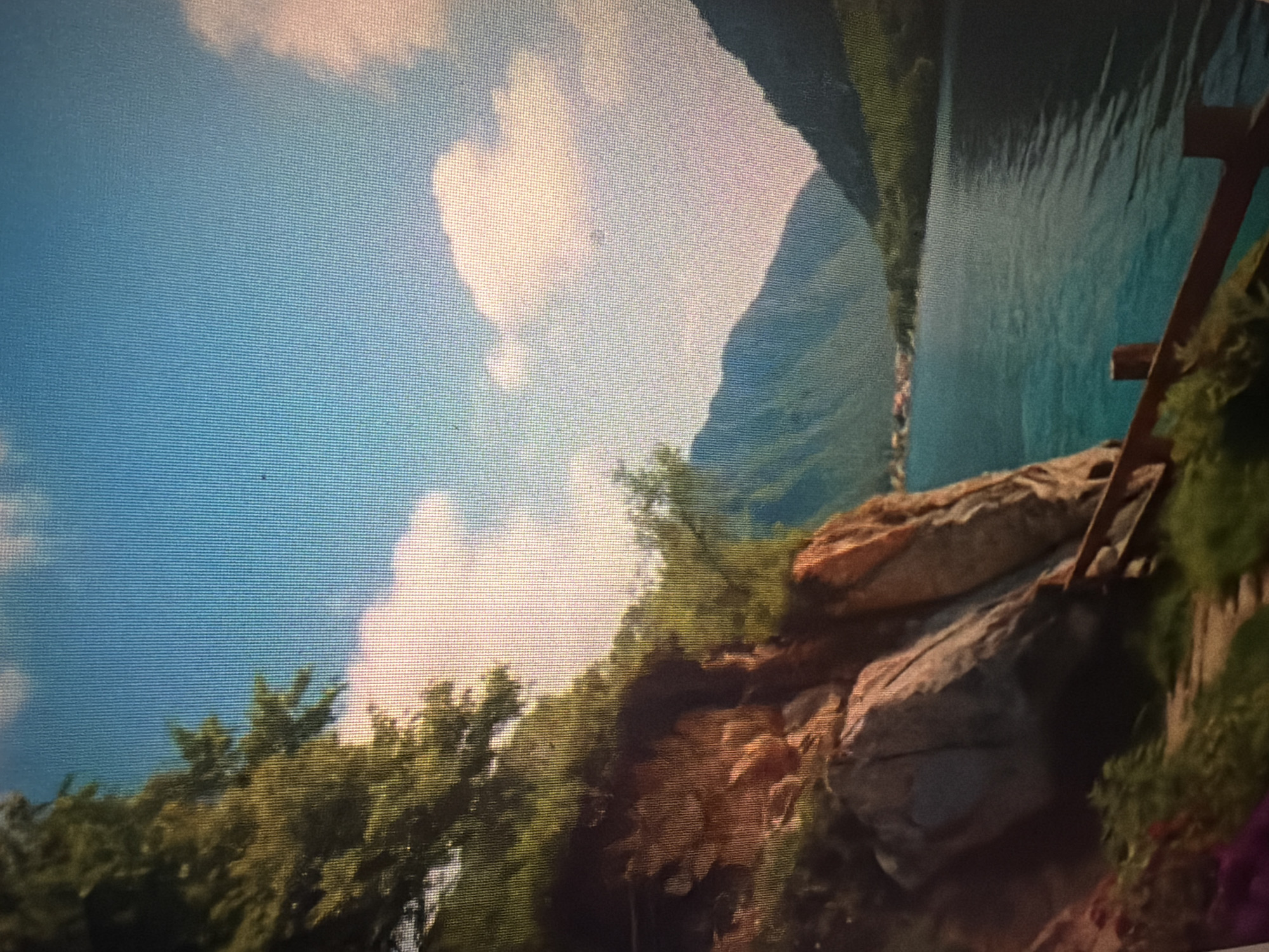 The image depicts a serene natural landscape. In the foreground, a rocky cliff face is covered with lush green foliage and trees. A wooden railing runs along the edge of the cliff, overlooking a tranquil body of water. The water appears to be a lake or a wide river, reflecting the clear blue sky and soft white clouds. In the distance, rolling mountains rise, their slopes covered in dense vegetation. A small cluster of buildings, suggesting a village, is visible on the far bank of the water, nestled amongst the trees. The overall impression is one of natural beauty and peacefulness. The image does not contain any people, animals, or any specific indication that it is located in Baan Mae Nam, Thailand.