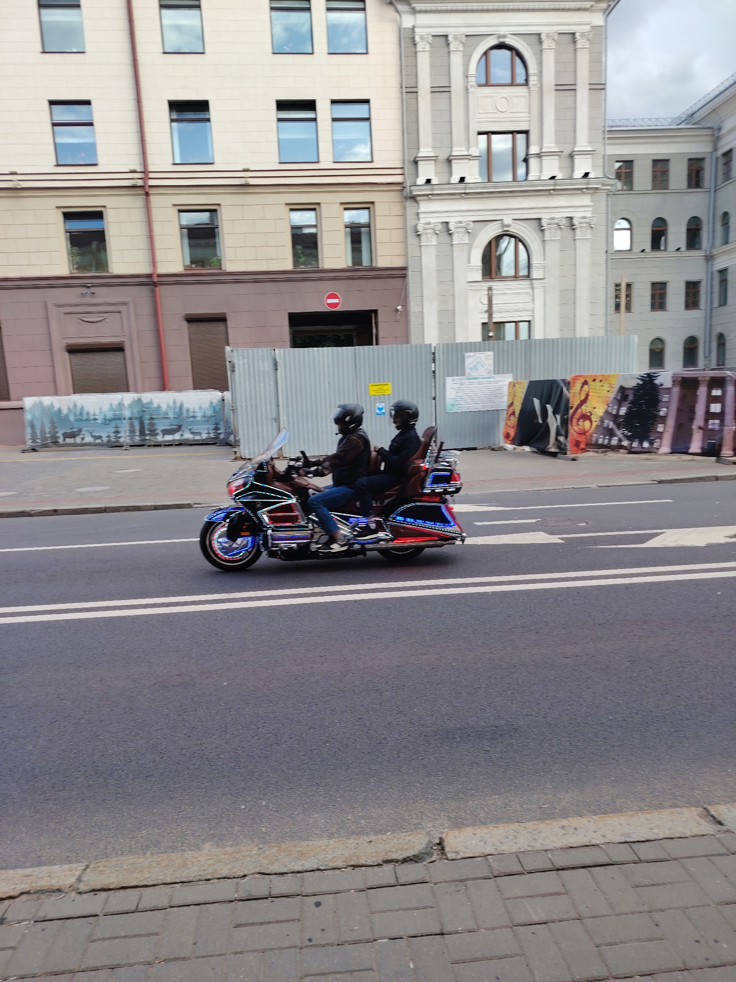 The image captures a street scene in the city center of Minsk, Belarus, likely along a major avenue such as Independence Avenue, characterized by its distinctive Neoclassical architecture. Two individuals, both wearing dark helmets and dark jackets with blue jeans, are riding a highly customized, large touring motorcycle. The motorcycle, possibly a Honda Gold Wing, is adorned with extensive blue, red, and white LED strip lighting along its frame, fairings, and wheels, creating a striking visual effect as it moves from left to right across the frame on a dark asphalt road.
The background features several buildings. On the left, a multi-story building with a light beige facade and numerous rectangular windows stands, featuring a reddish downpipe running vertically. Further right, a grand, ornate building in grey and white displays classical columns, decorative cornices, and arched windows on its upper floors, embodying a prominent Stalinist Empire style or Neoclassical architecture. To the far right, a simpler grey building with a mix of rectangular and arched windows is partially visible.
Along the sidewalk, a corrugated metal construction fence runs, partially obscuring the lower sections of the buildings. Sections of this fence are covered with murals: on the left, a forest scene with silhouetted deer against a blue-green background; on the right, a vibrant, multicolored mural depicting musical notes, a dark silhouette, and a stylized building cityscape. The street features clear double white lines separating traffic lanes and directional arrows painted on the asphalt. The sidewalk is paved with gray interlocking bricks. The sky is overcast, indicating daytime.