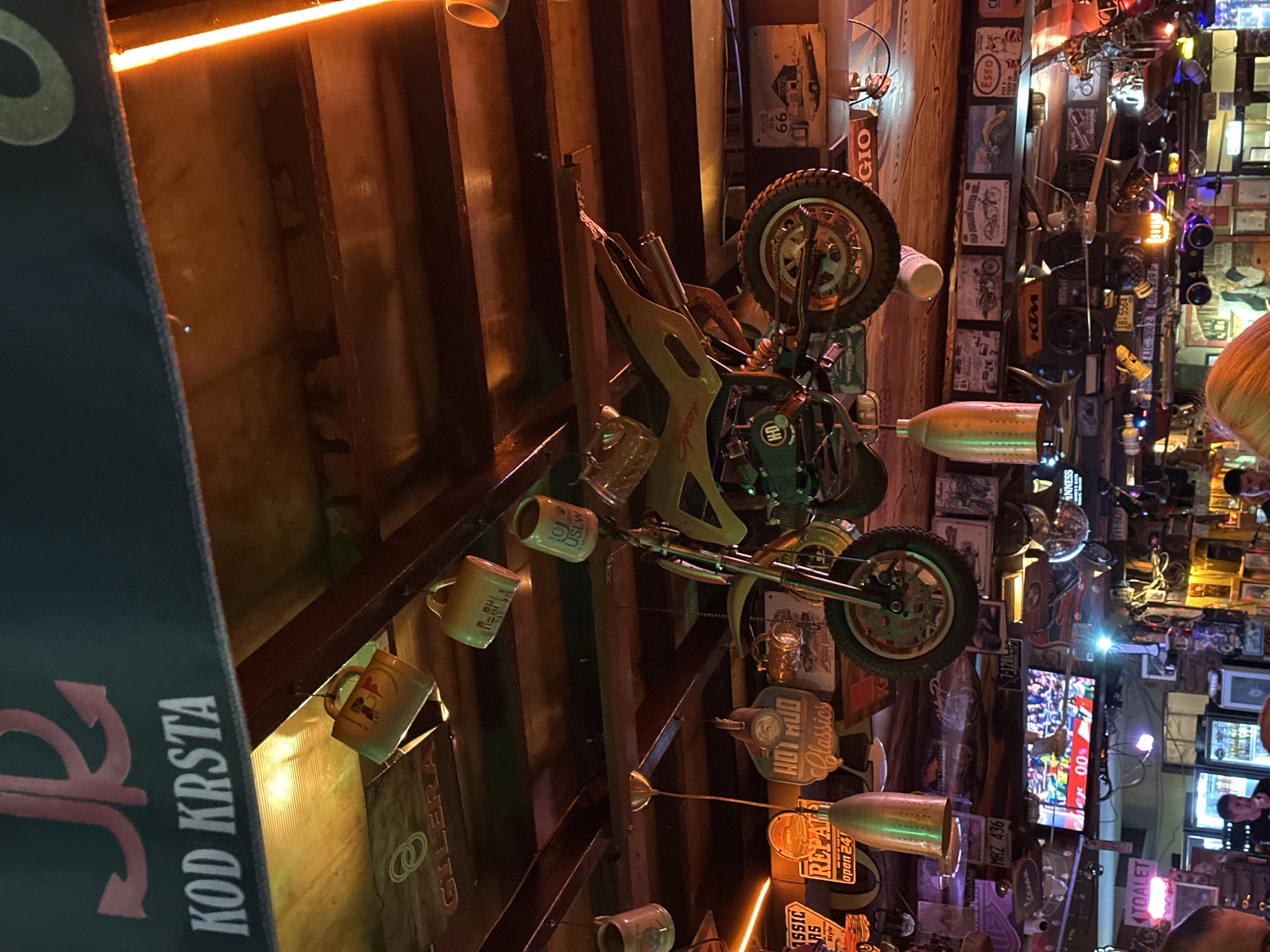 The image depicts the interior of a dimly lit bar or pub, likely in Budva, Montenegro, characterized by a rustic and eclectic decor. Hanging from the ceiling are several mugs, a miniature motorcycle, and various decorative items, including what appears to be a bullet-shaped pendant lamp. The ceiling is constructed of dark wooden beams, and overhead lighting casts a warm, orange glow.
Prominently displayed on a dark banner at the top of the image is white text reading "KOD KRSTA" and a stylized red anchor symbol. Below this, the walls and ceiling are adorned with numerous signs, license plates, and framed pictures, many with vintage or automotive themes. Signs like "HOT ROD Classics," "REPAIR," "ROUTE 66," and various brand logos such as "GILERA," "HD," "KTM," and "Esso" are visible. A small television screen is mounted on a wall, displaying an image of a sporting event.
Several individuals are present in the lower part of the image, mostly out of focus, suggesting the scene is a social gathering. The overall atmosphere is that of a themed establishment, likely catering to motorcycle enthusiasts or those who appreciate a retro aesthetic. The presence of the text "KOD KRSTA" and the anchor symbol may indicate a specific name or theme for the establishment.