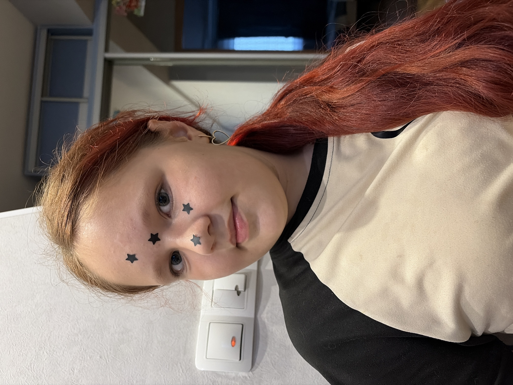 A young woman with light skin and red-toned hair is facing the camera. She has black star stickers on her face near her eyes and nose. She wears a black and white shirt. Behind her is a white wall with a light switch. A kitchen cabinet is also visible in the background. The location is Chaikovsky, Russia.