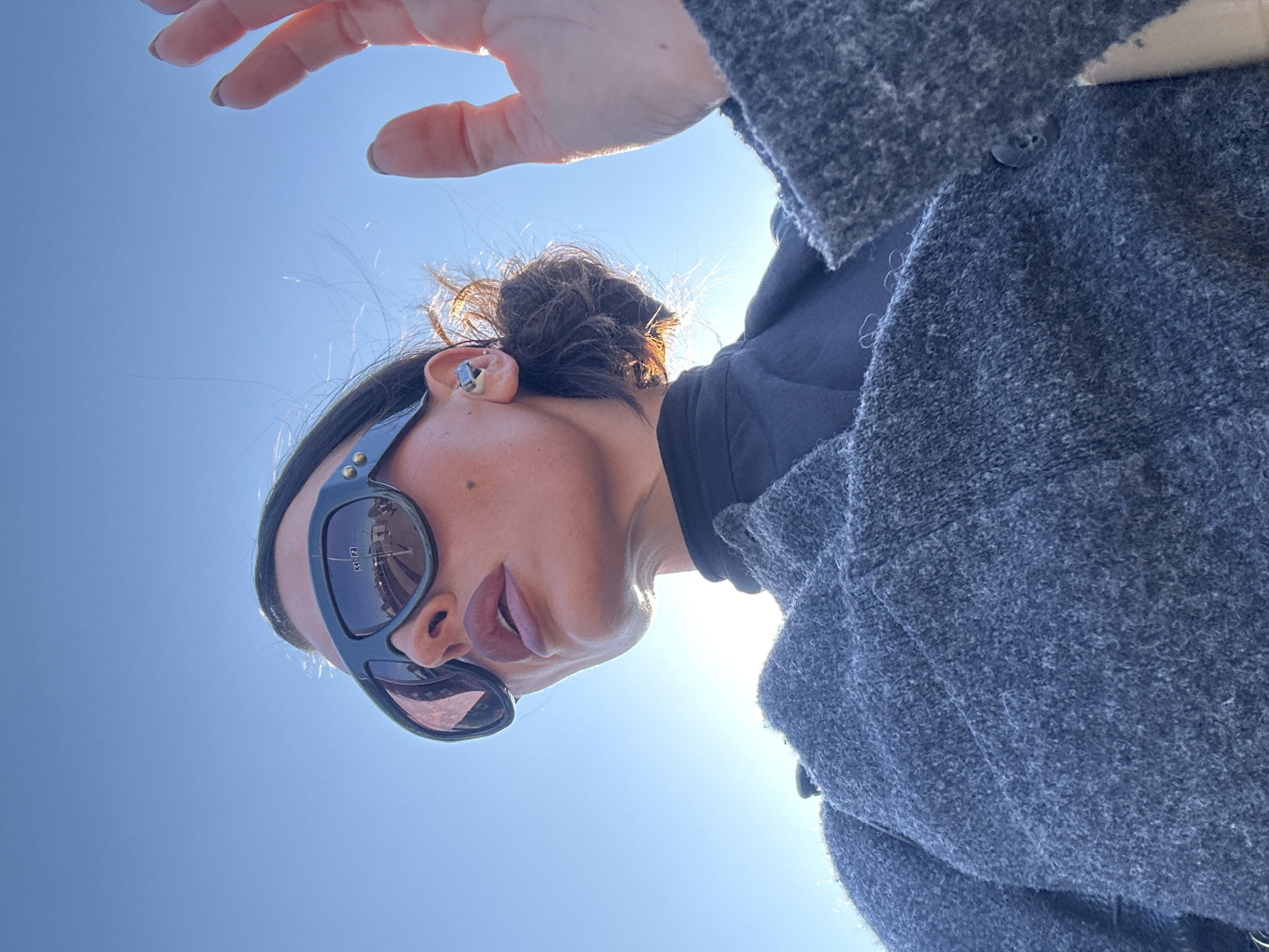 A low-angle shot captures a person, likely a woman, looking upwards against a bright, clear blue sky. Her head is angled to the right, and a bright sun flare emanates from behind her on the right side of the frame, illuminating the edges of her hair and coat. She wears dark, rectangular sunglasses that reflect a distorted view of what appear to be distant structures or scenery. Her lips are slightly parted, and she has an earbud in her visible right ear. Her dark hair is pulled back into a bun, with some stray strands catching the sunlight. She is dressed in a dark top and a textured, grey, possibly wool, coat. A hand, with lightly painted fingernails, extends into the top left corner of the frame, appearing to reach out or shield from the sun. The overall impression is a sunny, outdoor setting. There is no visible text.