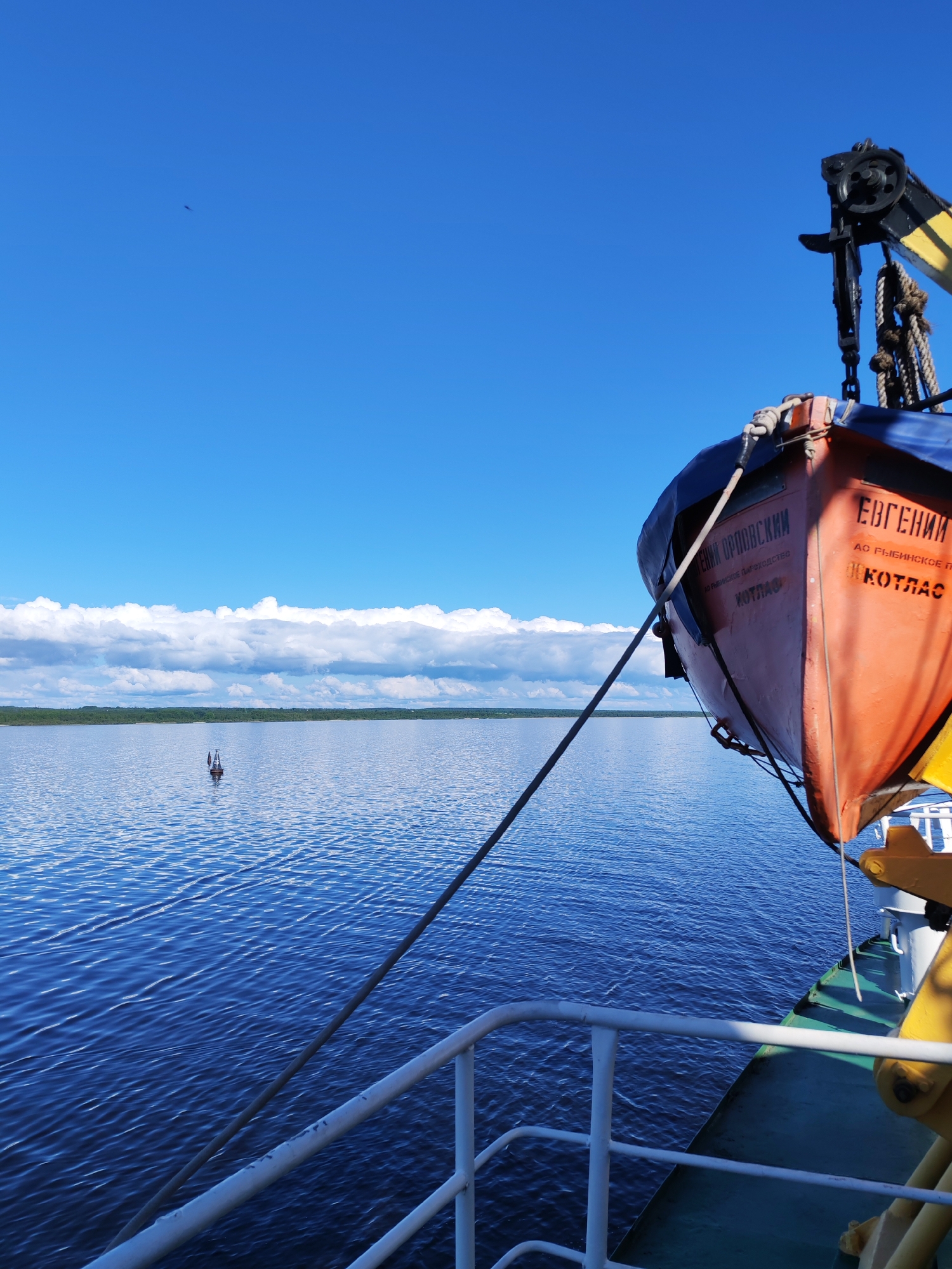The image depicts a view from a boat on a river or large body of water. A lifeboat, painted orange and blue, is secured on the boat's side. Text in Russian is visible on the lifeboat. A small navigation buoy marks the waterway. On the horizon, a line of trees borders the water, with a band of clouds in the sky above. The sky is largely clear and blue. The location might be near the city of Kotlas, as that is written on the lifeboat. Prislon' is not directly identified but could be nearby the area where this photo was taken. The general scene suggests calm weather and daytime.