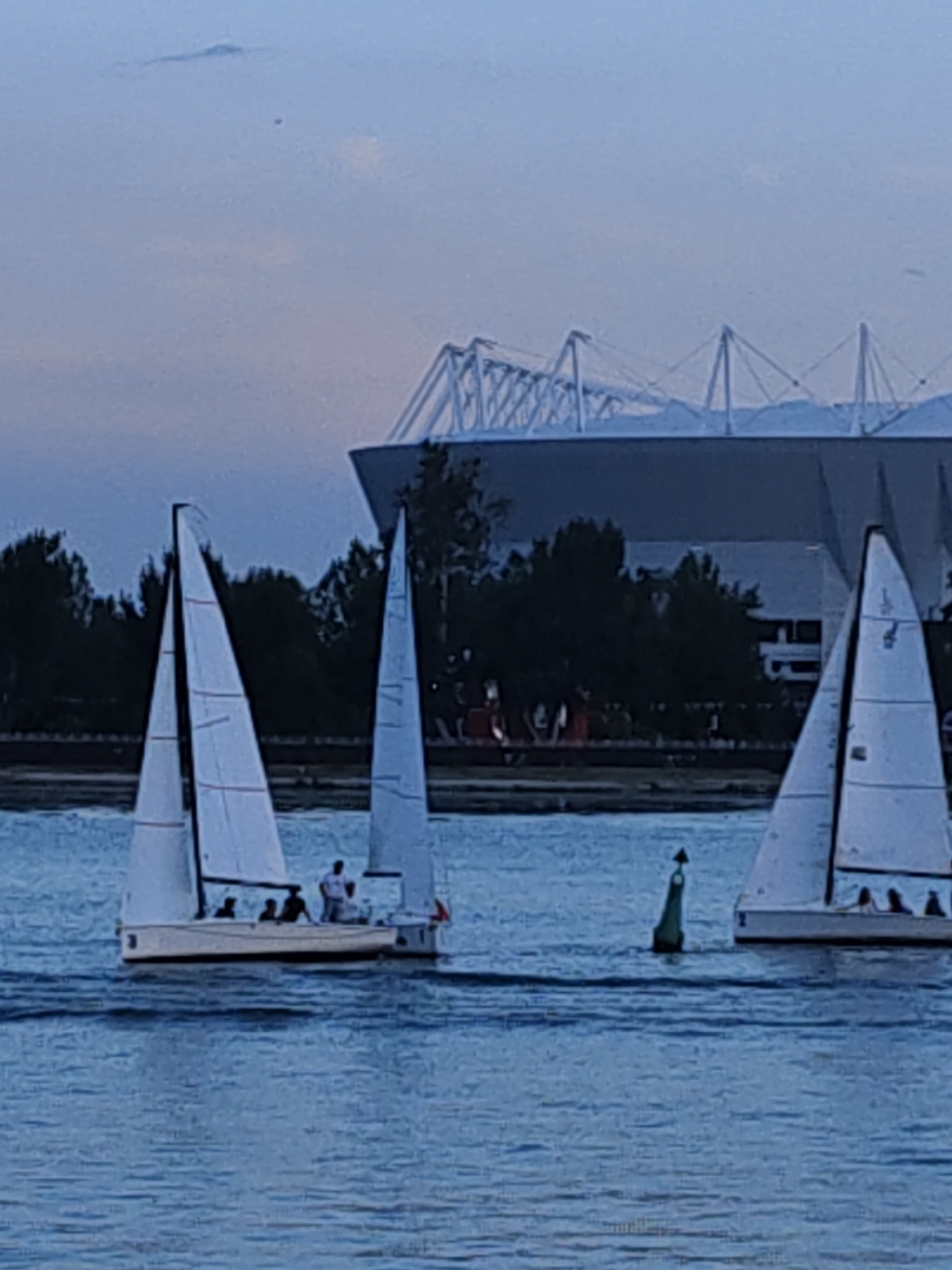 A nighttime photograph captures a body of water with several sailboats on it. In the foreground, a white sailboat with a red stripe on its sail is positioned to the left of the frame, its sail angled towards the right. A second white sailboat with a similar red stripe is to the right of the first, its sail also angled. A green buoy is visible in the water between the two sailboats. In the background, trees form a dark line across the horizon. Above the trees, a large, modern stadium structure with a white, skeletal roof is visible. The sky is a pale blue and appears to be twilight.