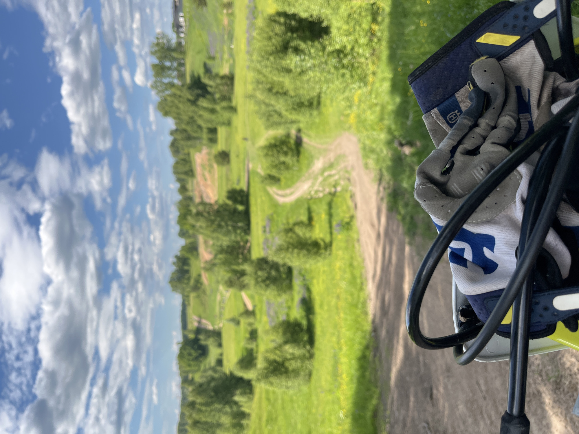 This image, captured in Новочувашский, Russia, presents a rural landscape from the perspective of an individual positioned on a dirt bike or similar off-road vehicle. In the immediate foreground, the black handlebars and control cables of a vehicle are prominently displayed. Beneath the handlebars, a white and blue motocross garment is visible, partially obscured by a pair of grey and black motocross gloves, which feature a visible Husqvarna logo on the wrist strap. The gloves are resting on the garment and a dark blue component of the vehicle's dashboard or front fairing.
The midground reveals a vast, undulating green landscape characterized by several unpaved dirt tracks winding through grassy fields. Numerous deciduous trees and bushes are scattered across the terrain, with some areas showing patches of purple wildflowers, likely lupines, amidst the green and occasional yellow flora. The dirt tracks suggest a prepared area for off-road riding.
In the background, the landscape continues with rolling green hills and a dense treeline under a bright blue sky. Numerous white, fluffy cumulus clouds are distributed across the sky. On the far right horizon, indistinct structures, possibly houses or buildings, are faintly visible. The scene is illuminated by strong daylight, indicating clear weather conditions. No individuals are visible in the frame.