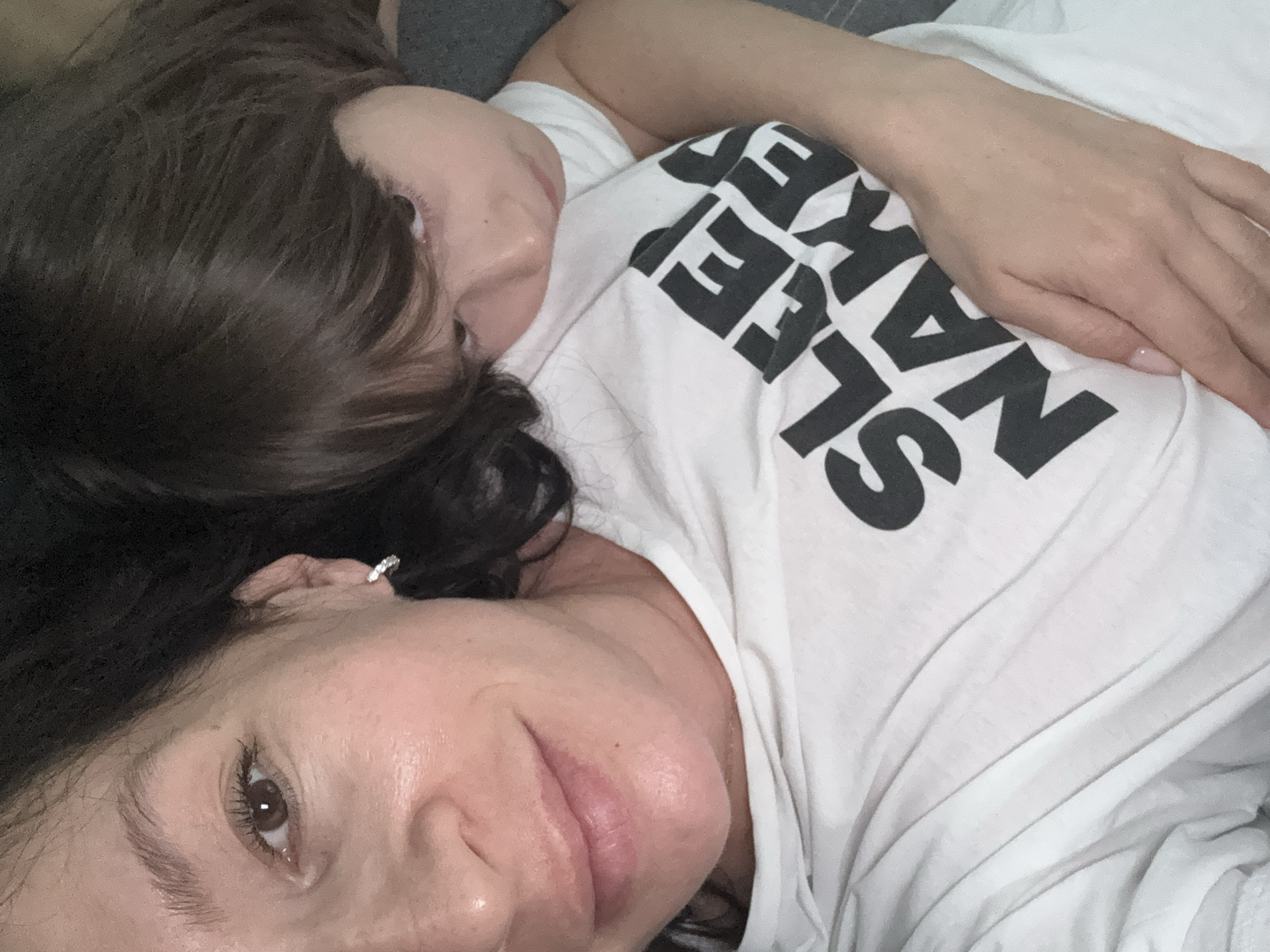 An intimate, close-up indoor shot features two individuals, likely an adult female and a child, lying down together. The adult female occupies the foreground, looking directly at the camera with a soft, content expression. She has dark hair and wears a white t-shirt with large, bold black text. Part of the text, "SLEEP NAKED", is clearly visible, with "SLEEP" appearing upside down relative to her head. Her right eye is visible, showing a brown iris and defined eyelashes, and a small, sparkling earring is seen on her right earlobe. Her lips are slightly parted, indicating a relaxed state.
Behind and slightly above the adult, the head of a child is visible, resting close to the adult's shoulder or chest. Only the child's dark hair and a small portion of their face are discernible, suggesting they are nestled against the adult. The setting appears to be a cozy domestic environment, likely a bed or couch, given the relaxed posture and close proximity of the individuals. The background is dark and out of focus, enhancing the focus on the subjects and their tender interaction. The overall mood of the image conveys warmth, comfort, and affection. The time of day is not explicitly indicated, but the scene suggests a moment of rest or bonding. The location context is Chaikovsky, Russia.