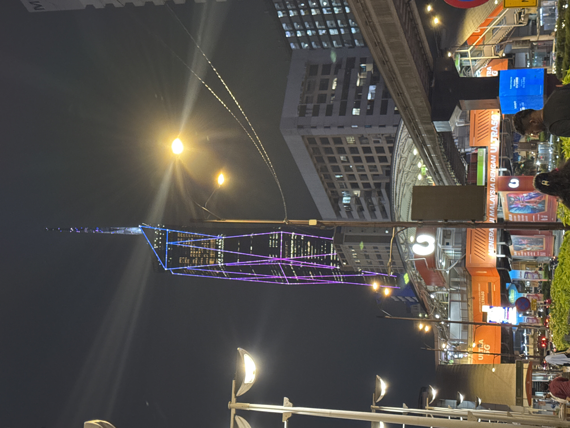 At night, the image shows a brightly lit cityscape. A tall skyscraper, adorned with geometric blue and purple lights, pierces the night sky. Streetlights illuminate the foreground. Buildings of varying heights and architecture are visible in the background. Commercial signage and advertisements are apparent, suggesting a bustling urban environment. People are seen walking. The image is set in Kuala Lumpur, Malaysia, and captures the city's modern urban landscape.
