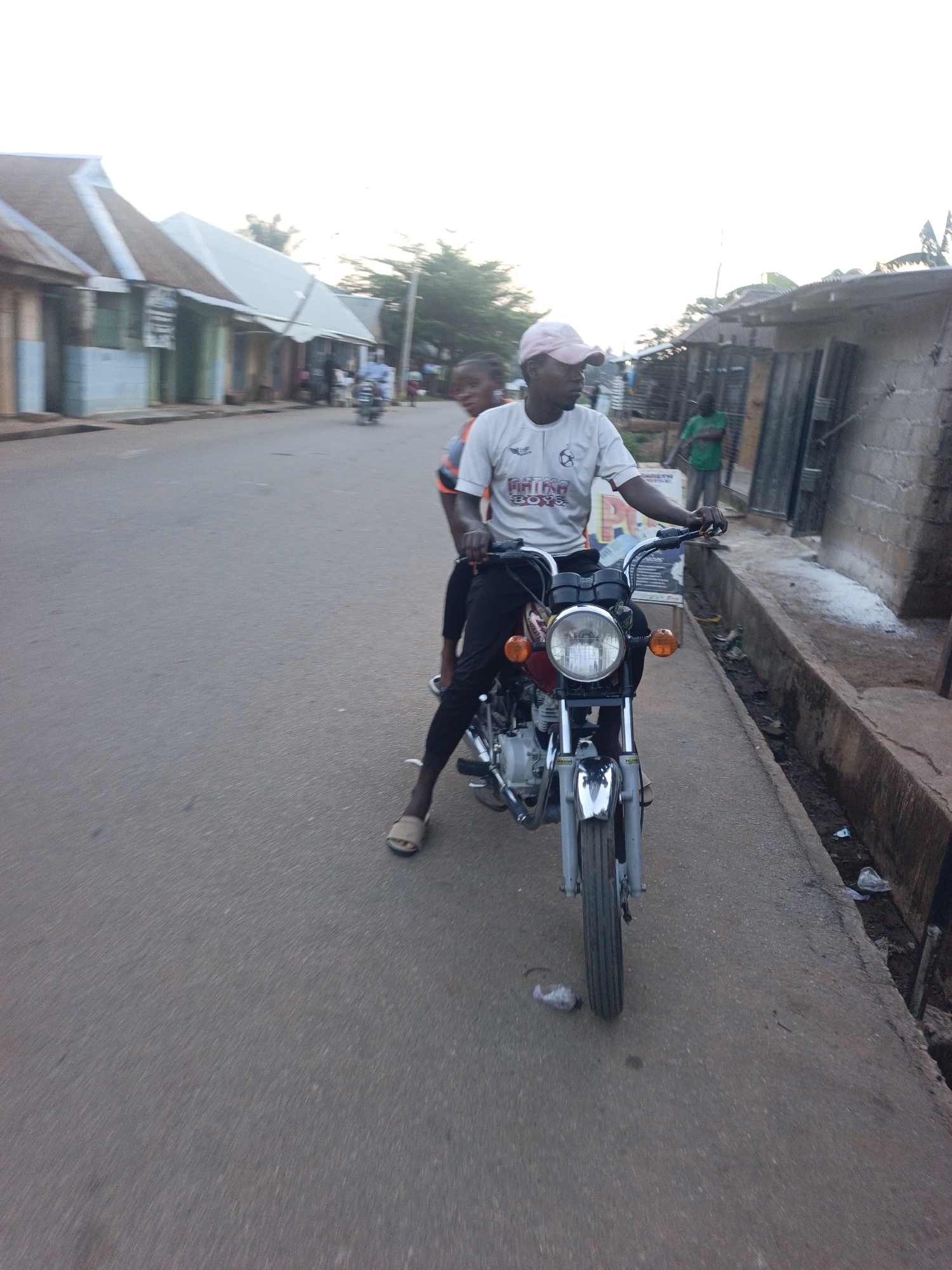 The image shows a man wearing a white t-shirt and a pink cap riding a motorcycle on a street in Akwanga, Nigeria. He has a passenger behind him. The street is paved with asphalt, and there are buildings lining the street on both sides. Some buildings appear to be houses or shops, and one has a corrugated metal roof. A man in a green shirt is standing near a building on the right. There is a small pile of debris near the curb. In the distance, more people and vehicles can be seen on the street. The sky is overcast, suggesting it may be early morning or late afternoon.