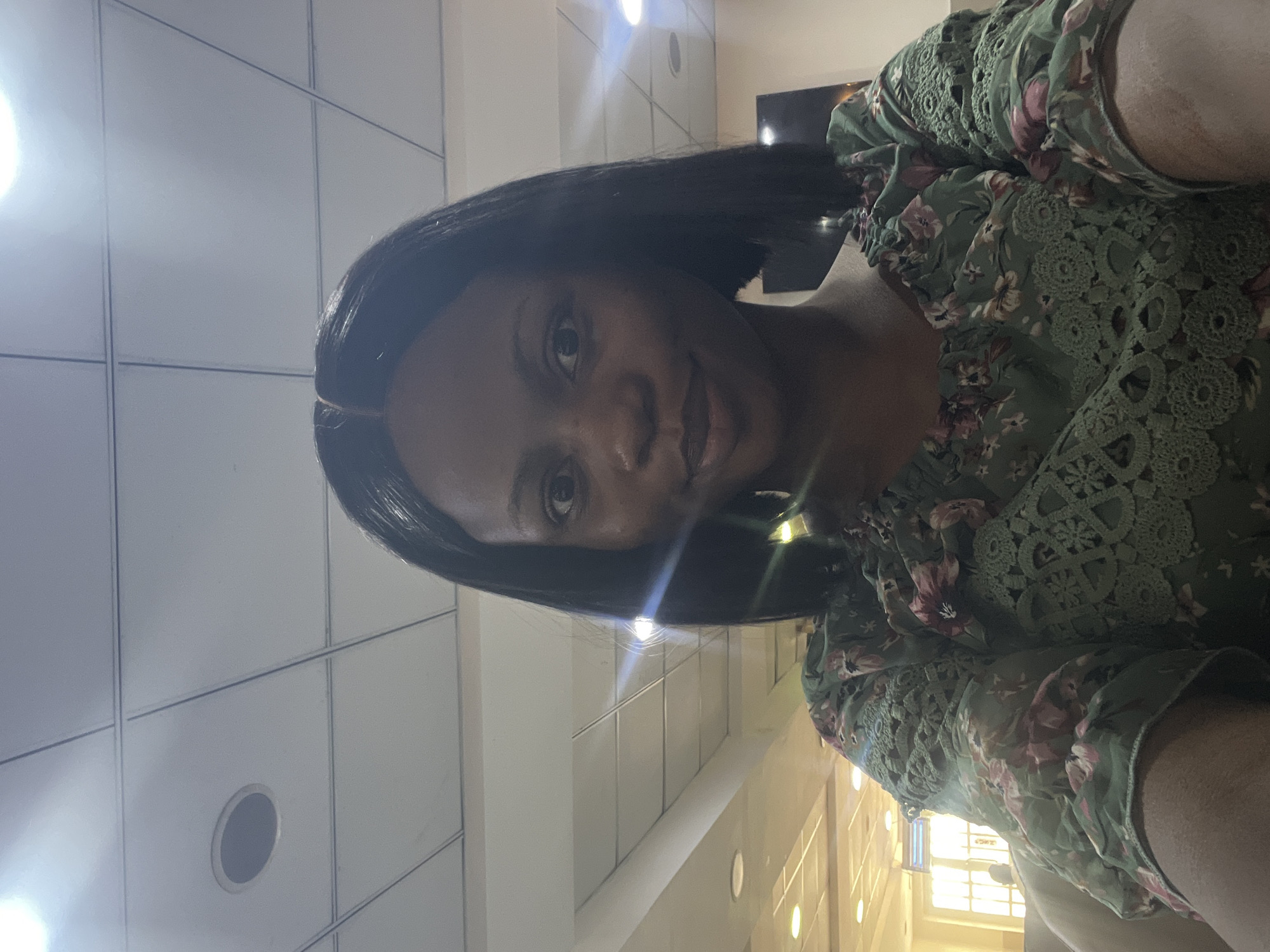 A medium close-up shows a woman with dark skin and short, dark hair. She is wearing a green floral top with lace details. The background appears to be an indoor space with a tiled ceiling and linear lighting, suggesting a commercial or public building. Given the location, it is plausible this is a business within Ikeja, Nigeria. No specific actions or interactions are visible in the image.
