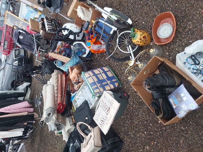 The image depicts an outdoor market stall in Eckbolsheim, France. The ground is covered with dark gravel. The scene is densely packed with various used goods for sale.
In the foreground, a brown cardboard box contains several pairs of dark boots and other packaged items. Adjacent to this box, a brass-colored chandelier with three frosted glass shades, a yellow ceramic jug with floral patterns, and a reddish-brown rectangular dish holding a clear, patterned glass plate are displayed on the ground.
To the left of the center, a large stack of rolled rugs and textiles, primarily in neutral and earth tones, rests on the ground. Nearby, a beige handbag, two black handbags, and a black backpack are placed. A white, flat object, possibly a book or sign with text and images, lies on top of a dark green container. A blue and white patterned tote bag is positioned next to it.
The mid-ground features a variety of items: a plastic doll with curly brown hair lying face-up amidst a collection of small objects, a light blue woven basket, an orange toy weapon, a white helmet, and various electronic components or small appliances. Further back, a dark-colored stroller is visible, along with a bicycle wheel and other mechanical parts. Two framed artworks, one a dark painting and another a lighter landscape print, lean against other items.
In the background, several vehicles are parked, including a dark grey Audi sedan with a partial French license plate (VV-435-FS) and a red vehicle. Clothing on hangers is visible on portable racks to the far left. No individuals are discernible within the frame interacting with the merchandise.