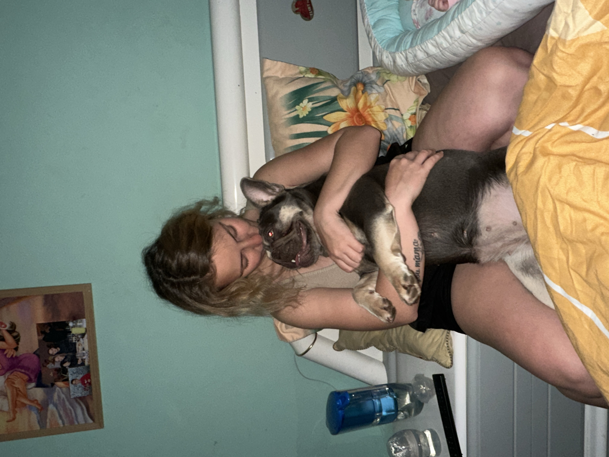 An indoor, dimly lit scene in Unknown, Unknown, shows a light-skinned female individual with blonde hair, visible from the chest down to her mid-thighs, seated on a bed. She is wearing a light-colored tank top and dark shorts. Her right arm displays a tattoo with script. She holds a brown and tan French Bulldog-type dog in her arms, pressing her head against the dog's head as if kissing it. The dog's mouth is slightly open, revealing its tongue, and its eyes exhibit red-eye from the camera flash.
To the left of the woman, on a light grey bedside table, are a blue and clear plastic water dispenser, a clear glass or cup containing ice, a black remote control, and a white lamp with a gold band. The wall behind her is painted a light teal color. A framed picture hangs on the wall, depicting two figures and a beach scene, with smaller framed photographs inset below it, showing a group of people and an individual in a red shirt.
Behind the woman, a white headboard or bed frame is visible. A floral pillow with yellow and green designs rests against the headboard. To the right of the woman, a baby's hand is visible within a light blue and white bassinet or co-sleeper. The bed is covered by a grey blanket, partially obscured by a yellow comforter with white stripes. A red, heart-shaped object with the word "FLIRT" is affixed to the white headboard.