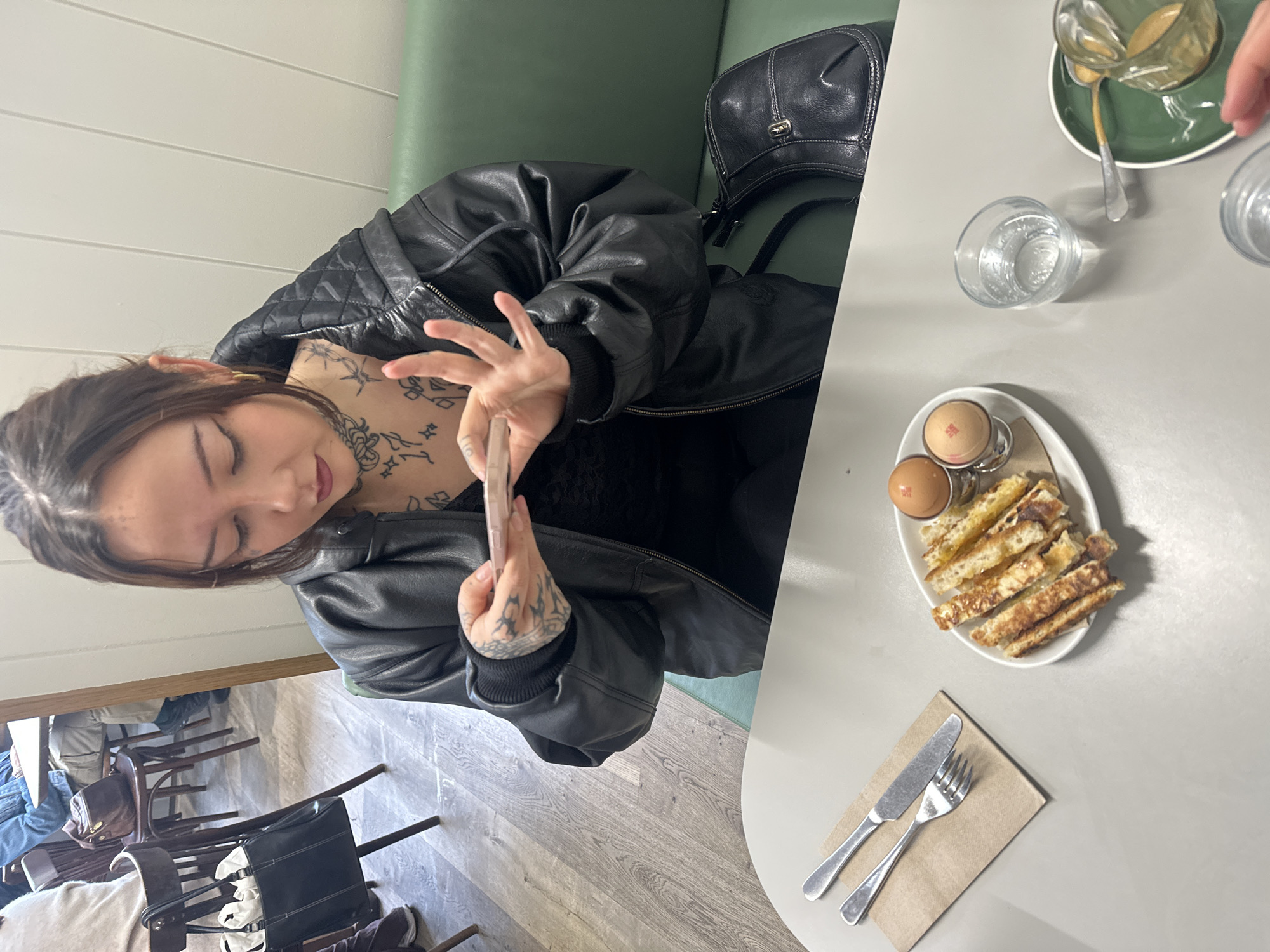 A woman with dark hair and numerous tattoos is seated at a table in what appears to be a café or restaurant. She is wearing a black leather jacket and holding a mobile phone, possibly taking a picture. A black handbag is visible beside her. The table is set with breakfast items, including two boiled eggs in egg cups, strips of toast, a glass of water, and a cup of coffee. Cutlery is laid on a napkin. The interior of the location has light grey walls and what appears to be light green upholstered seating. Other tables and chairs can be seen in the background. Given the style and location of the subjects, this appears to be a local business.