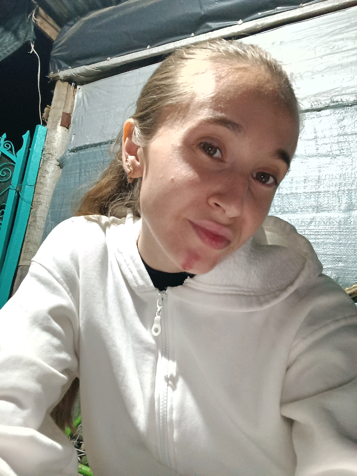 A close-up, waist-up selfie shows a young woman with blonde hair pulled back. She is wearing a white hooded sweatshirt with a zipper. She has brown eyes and is looking directly at the camera. A small red mark is visible on her chin. The background is out of focus, but appears to be an outdoor setting with a corrugated metal wall and some green fencing. The image was taken in Vypasne, Ukraine.