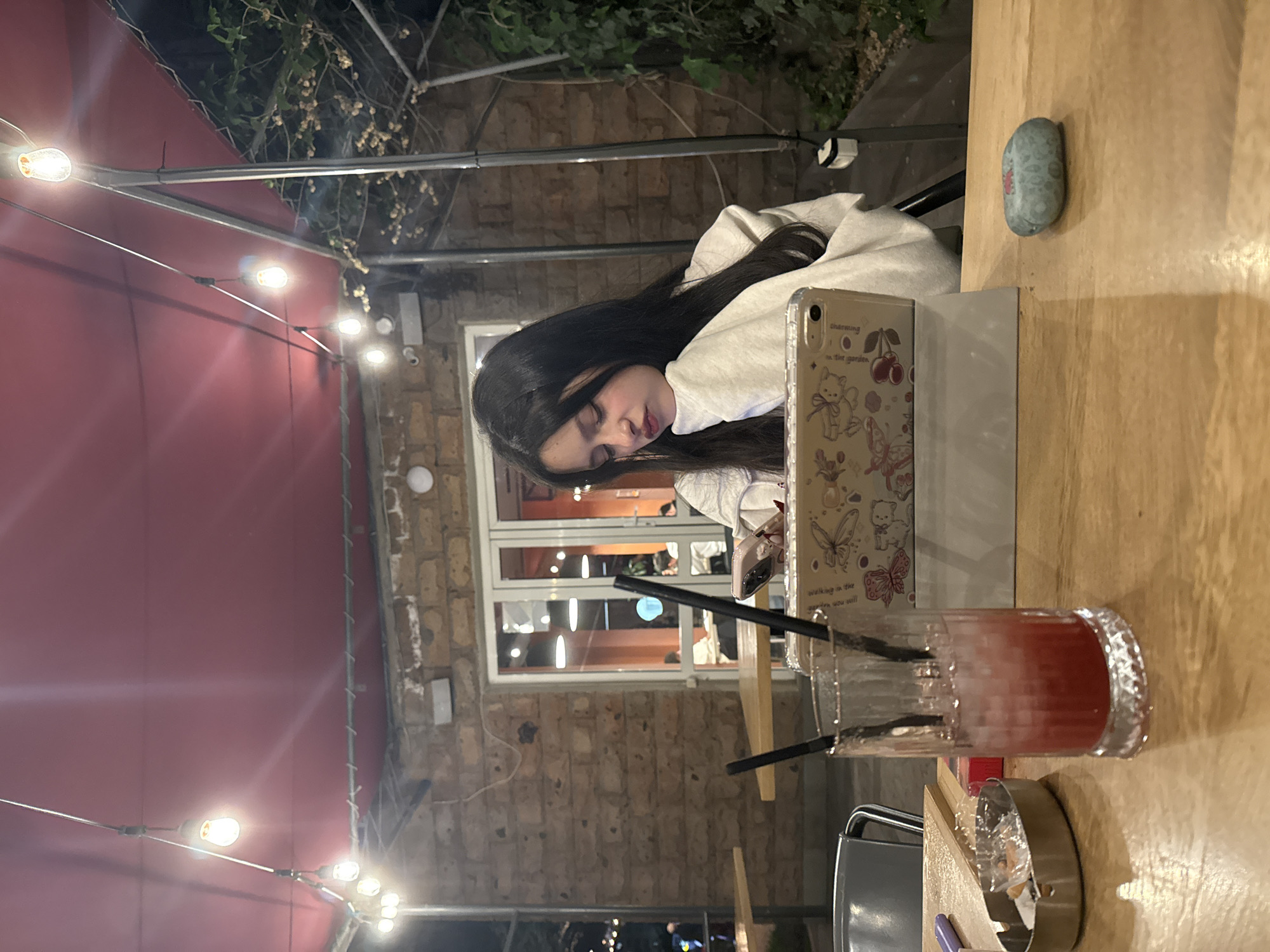 The scene is an outdoor dining setting in Yerevan, Armenia, at night. A woman with dark hair is resting at a wooden table. On the table are a phone with a decorated case, a glass of a red drink, an ashtray, and a blue object. The background features a brick wall, a window, and outdoor lighting. A red awning and greenery are visible above.