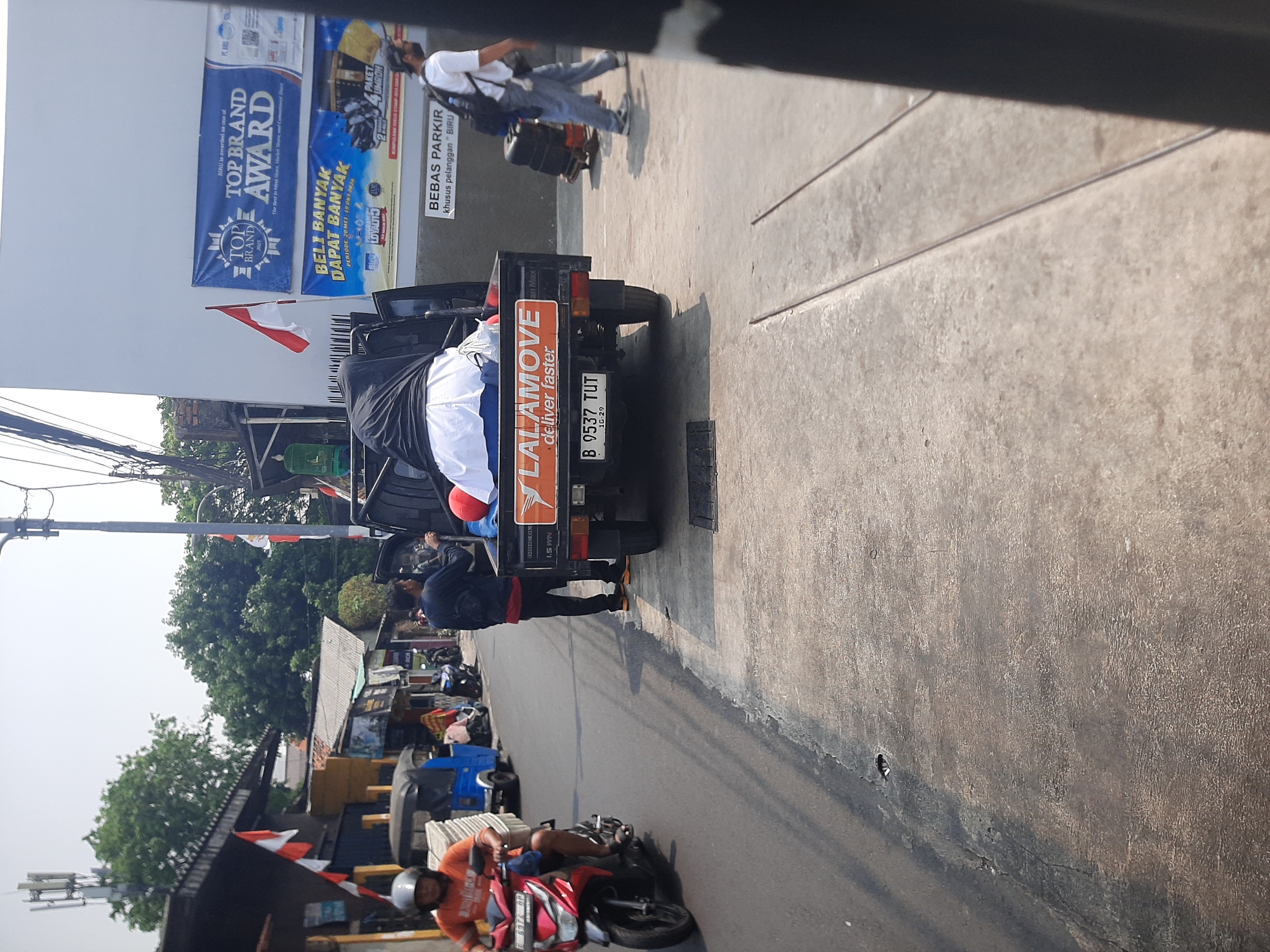 The image depicts a street scene in Central Jakarta, Indonesia, with a focus on a pickup truck and its surroundings. A red motorcycle is visible on the left, ridden by a person wearing an orange shirt and a helmet. The truck, positioned in the center, is loaded with bundled items covered in white fabric and a black cloth. The rear of the truck features a "Lalamove" logo and a license plate reading "B 9537 TUT 10-29". Two men are standing near the truck; one in a dark jacket and red shirt, and another wearing a white t-shirt and a backpack, pulling a rolling suitcase. In the background, buildings with advertisements, including a "TOP BRAND AWARD" banner, line the street, along with trees and a cell tower. The ground is paved with concrete.