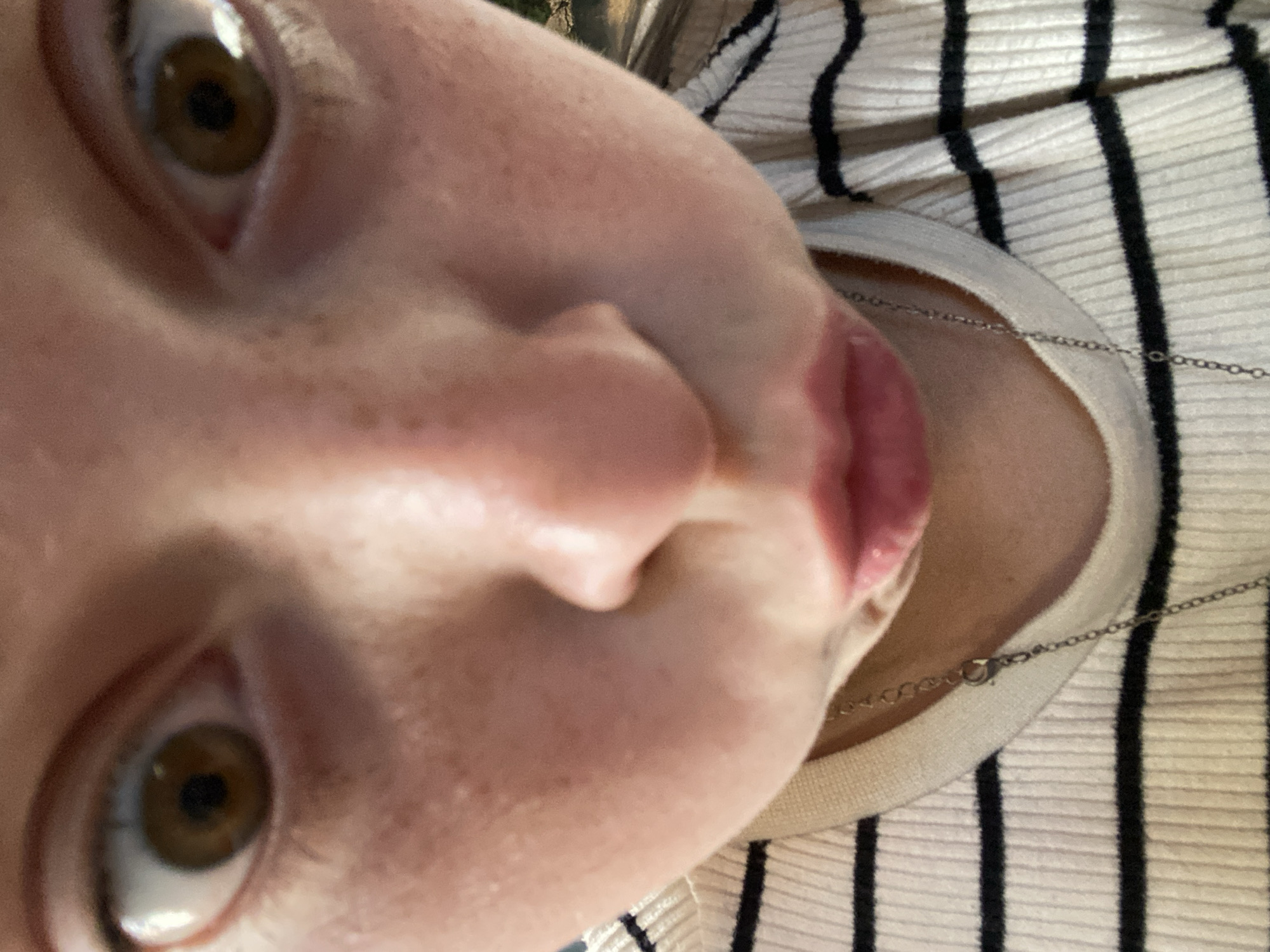 The image is an extreme close-up, low-angle, upside-down shot of a person's face, with the camera positioned very close to their nose and mouth. The subject has fair skin with visible freckles and brown eyes. Their mouth is slightly open, with the lower lip and tongue visible. A thin silver chain necklace rests on the person's chest, over a ribbed, cream-colored shirt with black stripes. The background shows a hint of trees and a bright sky, suggesting the photo was taken outdoors during the daytime. No text is visible in the image.