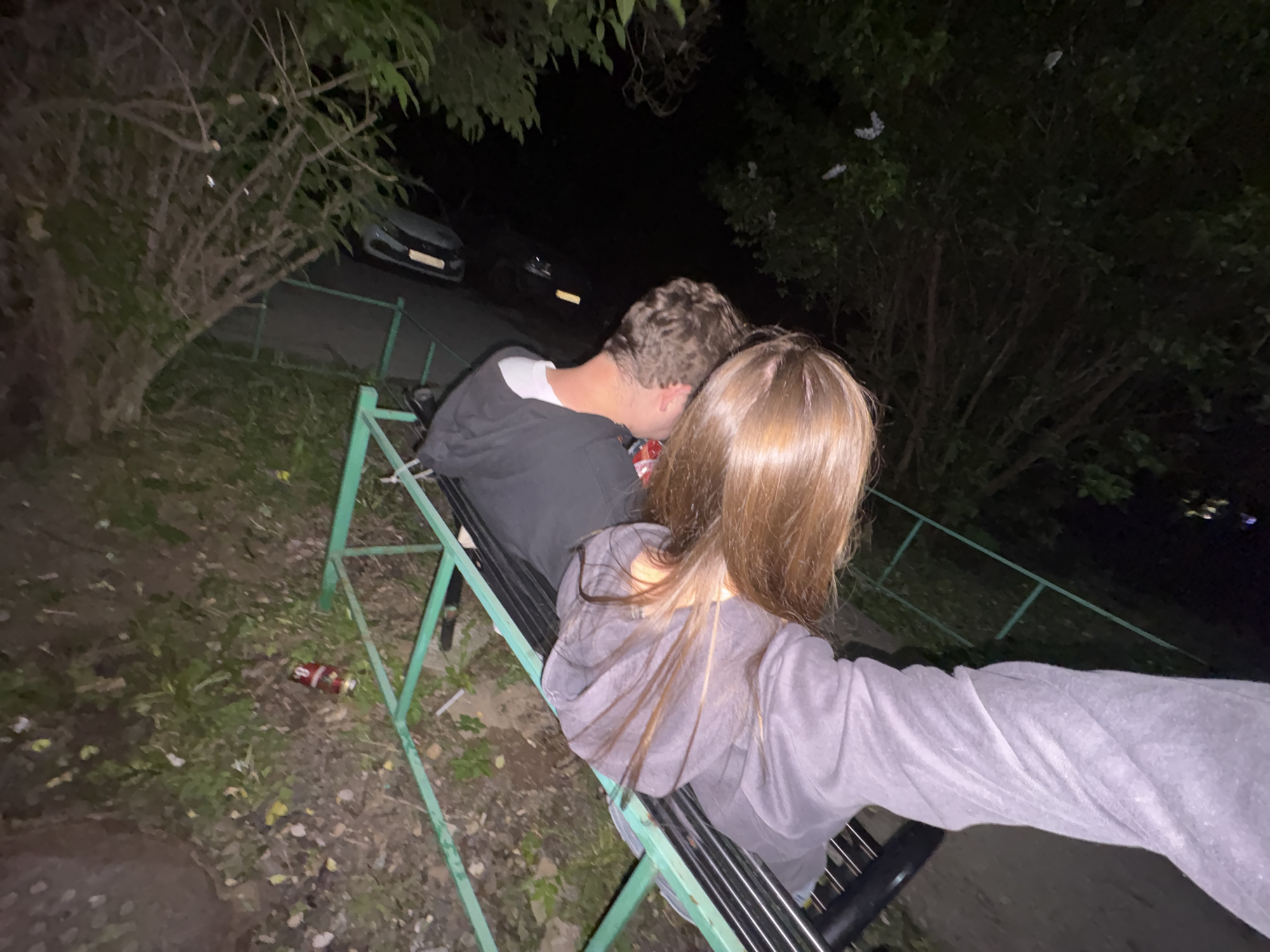A low-angle shot captures two individuals, one with long brown hair facing away and another with short curly hair, sitting on a green metal public bench. The picture seems to be a selfie taken by the person with long hair, as their arm is visible stretching out towards the camera. The scene is outdoors at night, with trees and shrubs bordering the area. In the background, there is a street with at least two cars parked along the curb. A can is visible on the ground to the left of the bench. The overall ambiance suggests a park-like setting. This scene is potentially in Perm, Russia.