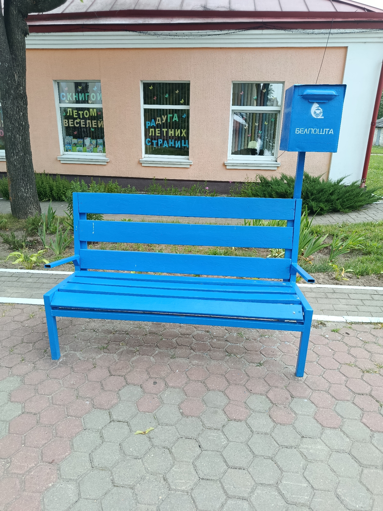A bright blue wooden bench and a blue mailbox are situated on a hexagonal paver stone pathway in front of a light peach-colored building in Zhlobin, Belarus. The building has two windows with decorative lettering and butterfly cutouts visible. The window on the left has text that reads "С КНИГОЙ ЛЕТОМ ВЕСЕЛЕЙ," and the window on the right has text that reads "РАДУГА ЛЕТНИХ СТРАНИЦ." The mailbox displays the logo of "БЕЛПОШТА." A tree trunk is visible on the left side of the frame, and green bushes and grass surround the area. The overall scene appears to be an outdoor public space.