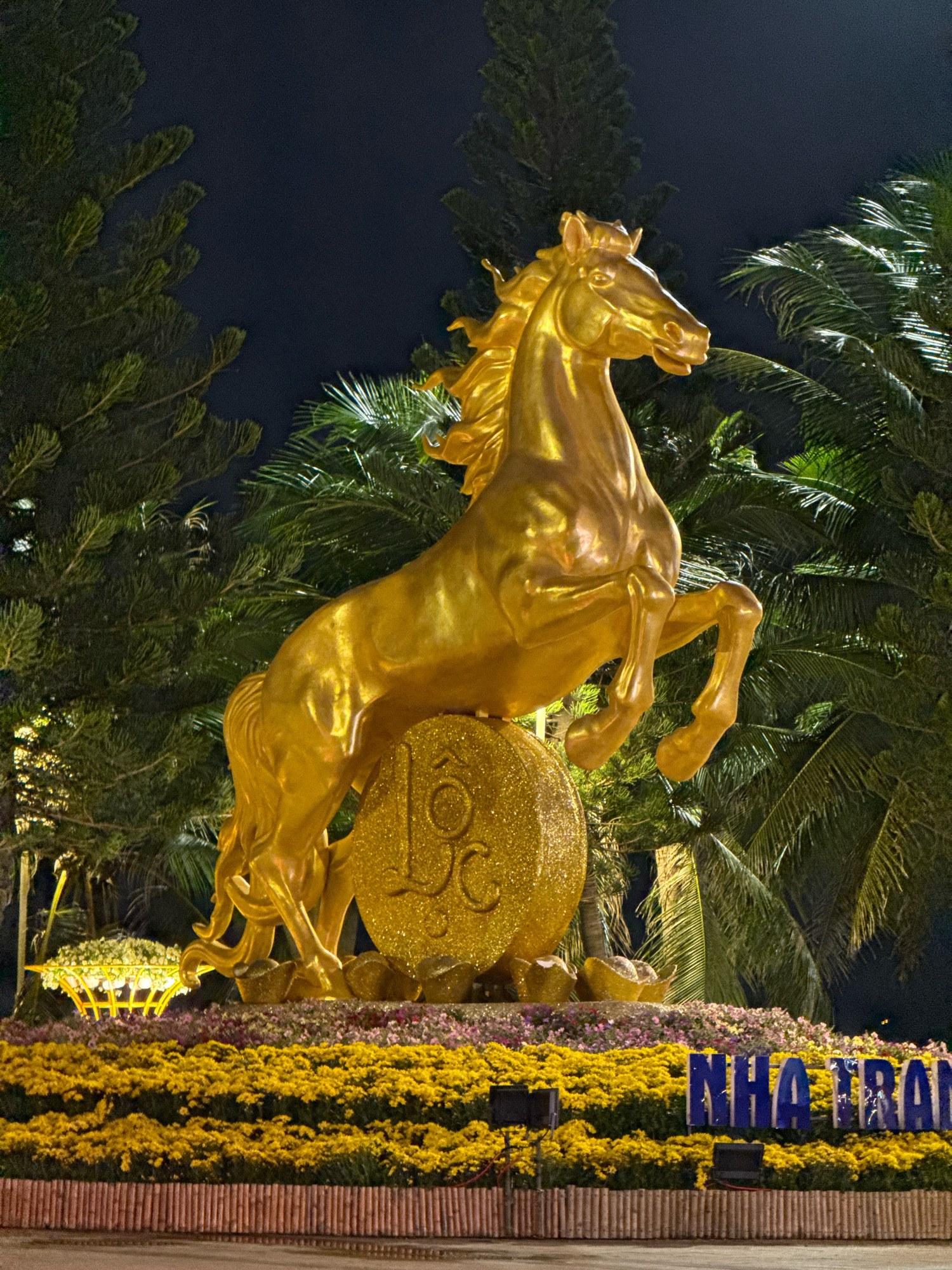 A gilded statue of a horse rears on its hind legs, its front hooves raised, atop a large, sparkling golden coin bearing the characters "18C." The horse is positioned in the foreground of the image, with lush green palm trees and pine trees forming a dark backdrop against a night sky. Below the statue, a vibrant display of yellow and pink flowers is arranged in a tiered fashion. To the right of the flowers, a blue sign with white lettering reads "NHA TRANG." The scene appears to be an outdoor decoration, likely for a festival or public event. The overall impression is one of grandeur and festivity.