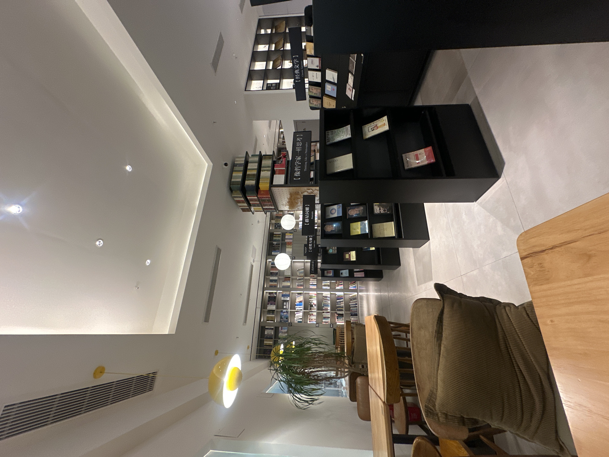 This image captures the interior of a modern bookstore or book-themed cafe located in Chongqing High-tech Zone, China. The space features a clean, contemporary design with light-colored flooring and white walls, creating a bright and airy atmosphere under ample artificial lighting, including recessed spotlights, linear strips, and distinct pendant lights like white spheres and a prominent yellow, egg-yolk-shaped fixture.
Tall bookshelves line the walls, filled with various books, while black display stands are strategically placed in the foreground and center, showcasing individual titles. A seating area with light-colored wooden tables and chairs, some fitted with olive-green corduroy cushions, occupies the right side of the frame, suggesting a comfortable spot for reading or relaxation. A large potted plant provides a touch of nature. Air vents are visible on the ceiling and walls.
Several signs with both Chinese characters and some English translations categorize the book sections. Visible text includes: "【群星闪耀】" (Stars Shine Brightly), "【诺奖专题】" (Nobel Prize Special), "【像哲学家一样思考】" (Thinking Like a Philosopher) accompanied by its English translation, and "【经典文学】" (Classic Literature). Part of "【我的职业】" (My Profession) is also discernible. No people are present, lending a serene and quiet ambiance to the well-organized and inviting environment.