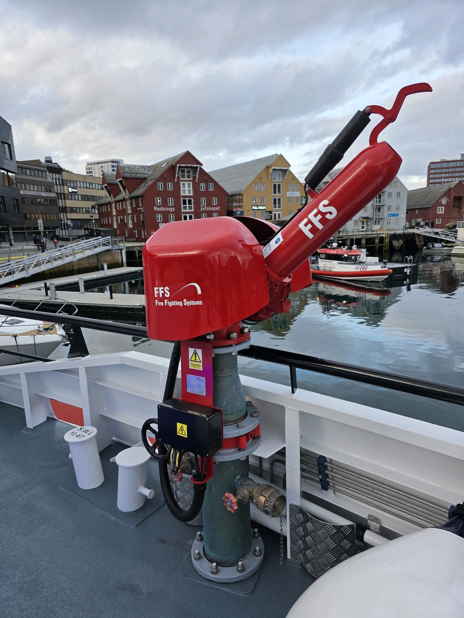 The image shows a red FFS Fire Fighting Systems monitor mounted on the deck of a vessel in a harbor. The monitor is a large, red, mechanical device with a nozzle at the top and a control arm. It is positioned on a green pipe that is bolted to the deck. The vessel's deck is painted gray and has several white structures and a black railing. In the background, there are several buildings with red and yellow facades, typical of Tromsø's harbor area, along with a calm body of water reflecting the overcast sky and the surrounding architecture. Several boats are docked in the harbor, including a red and white SAR (Search and Rescue) vessel. The overall scene suggests a functional, industrial environment within a city.