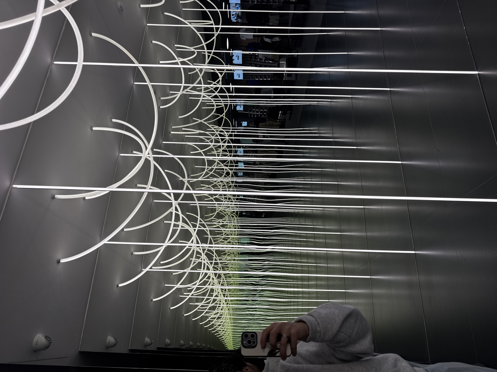 The image shows an interior space featuring an infinity mirror effect created by parallel walls adorned with horizontal linear light fixtures. Interspersed among the straight lights are curved lighting elements that resemble arcs, creating a repeating pattern that diminishes into the distance. The reflection of the Puma logo on screens is visible along the central axis, suggesting that this space is inside a Puma retail store. A person holding a smartphone is visible in the foreground, capturing the mirrored effect. The camera is focused, as seen by the visible reflection, and their arm is dressed in a gray long-sleeved sweatshirt. The location is inferred to be Yerevan, Armenia, presumably within a Puma store featuring this design element. Several security cameras are also visible.