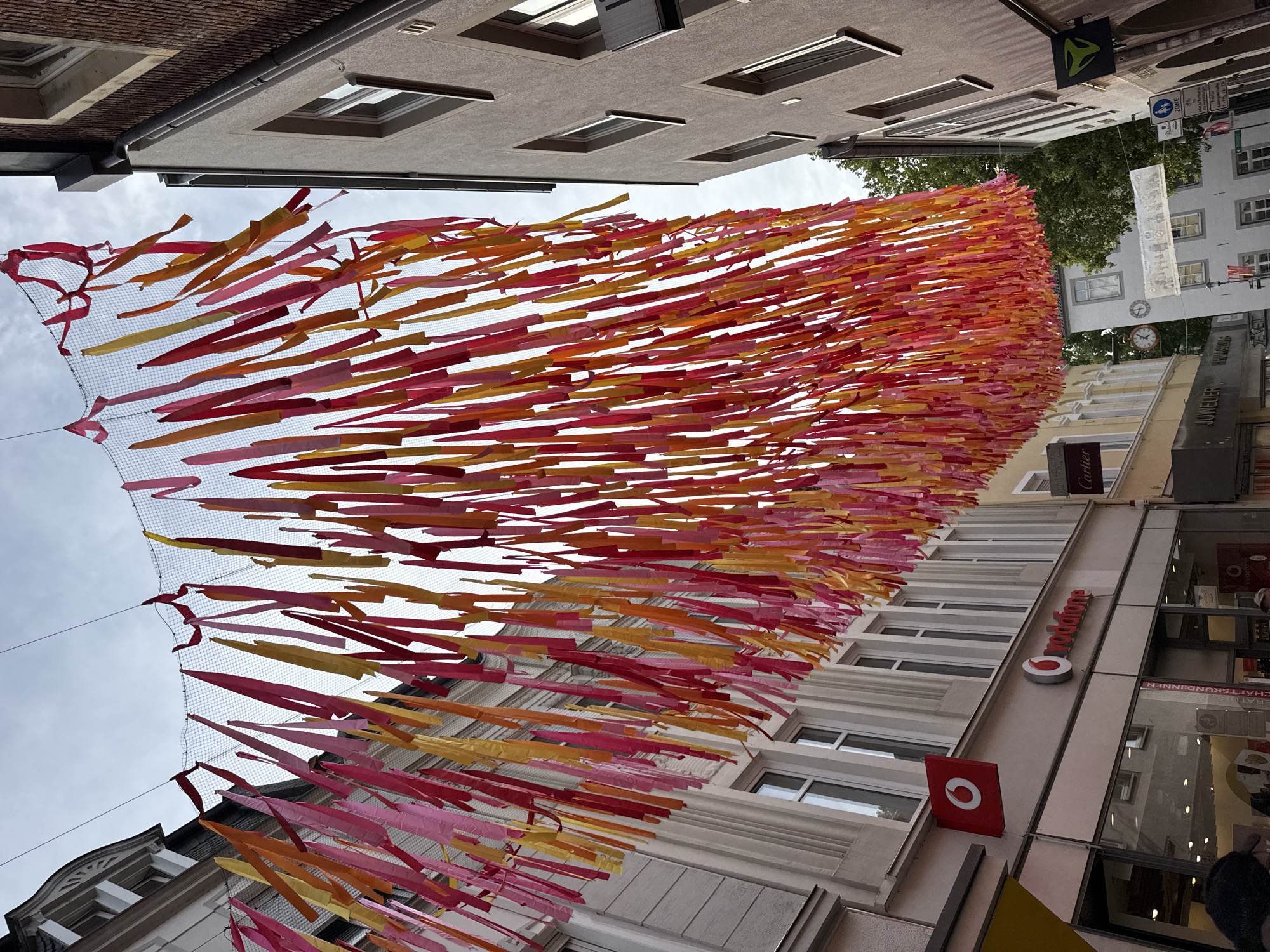 The image shows a street scene in Rheine, Germany, likely in the city center or a pedestrian shopping area. The focus is on a vibrant display of colorful fabric streamers, primarily in shades of pink, orange, and yellow, suspended across the street from a mesh netting. These streamers hang down, creating a festive atmosphere above the buildings.
On the left side of the street, a building with a white facade features multiple windows and a prominent red sign with a white logo that reads "Vodafone". Below this sign is a storefront, also labeled "Vodafone", with a large glass window showcasing the interior. Further down the street, a sign for "Cartier" is visible.
On the right side of the street, a darker building with brick details and windows is visible. A sign with a green logo is mounted on this building. Further down, more buildings are visible, some with arched doorways and windows. A street clock and several traffic signs, including a pedestrian crossing sign, are also present.
The sky above is overcast. The perspective is from ground level looking upwards towards the streamers and the buildings. No people are clearly identifiable or engaged in any specific actions in the foreground, though some blurry figures may be present in the lower part of the image. The overall impression is a cheerful and decorated urban environment.