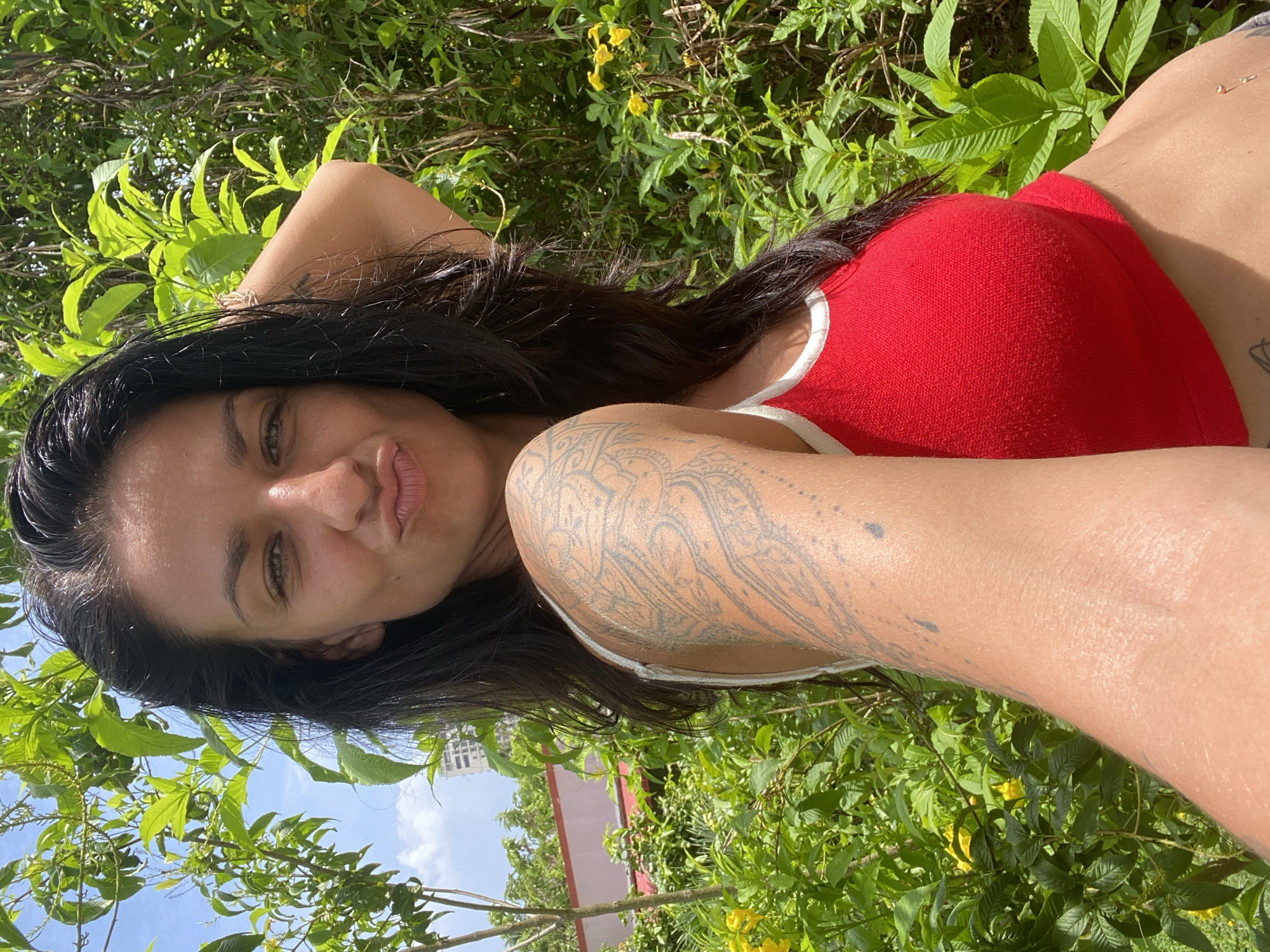 A woman with dark hair and green eyes is taking a selfie outdoors. She is wearing a red tank top with white trim. Her left arm is raised, and her hand is resting on the back of her head. A large tattoo is visible on her left arm. She is looking at the camera and puckering her lips. She is surrounded by lush green foliage, and the sky is visible above. In the background, a building and some trees can be seen. The location appears to be a tropical or subtropical environment, consistent with Mũi Né, Vietnam.