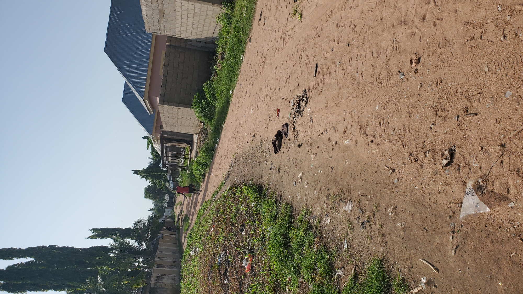 The image shows a dusty, unpaved road in Jalingo, Nigeria, with buildings under construction and tall trees on the left. A young person wearing a red shirt and dark shorts is walking away from the camera down the road. The roadside on the left is overgrown with grass and littered with debris, including plastic bags and other refuse. On the right side of the road, several buildings are in various stages of construction, with concrete blocks forming walls. Some dark-colored footwear lies on the dirt road. The sky is clear and bright.