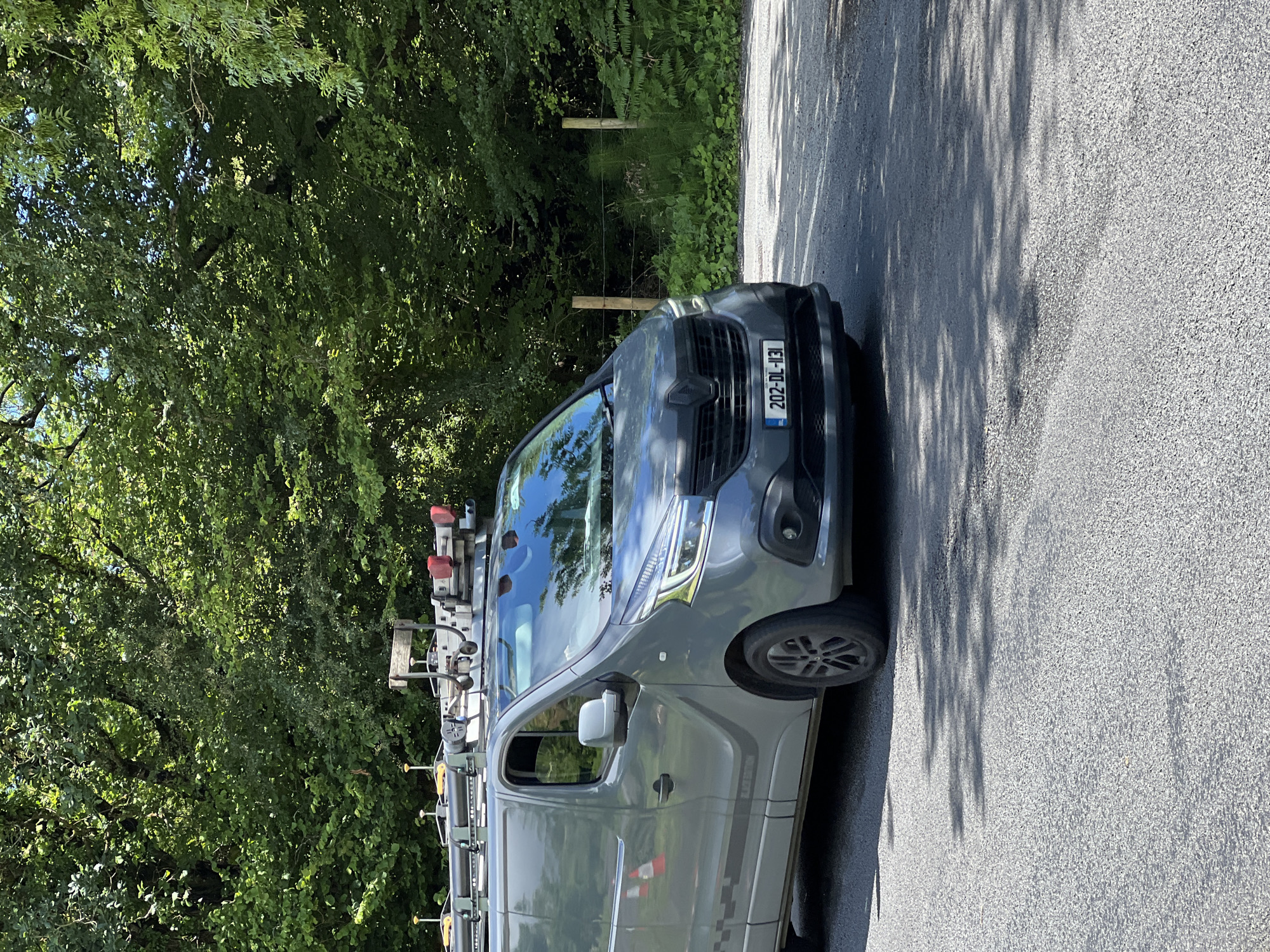 This media file displays a gray van parked on an asphalt road next to a dense green tree line. The van appears to be a work vehicle, equipped with a ladder rack on top and what looks like professional equipment attached. A fence, made of wooden posts and wire, separates the road from the wooded area. The license plate on the front of the van reads "202-DL-131". The scene is illuminated by bright sunlight, casting dappled shadows from the trees onto the road, suggesting it is daytime. The overall impression is a rural or suburban setting, possibly in Mountcharles, Ireland, given the location context. There are no people visible in the frame.