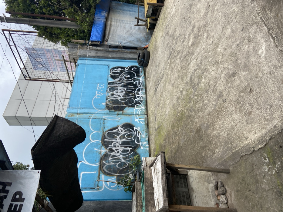 A large, blue, metal gate is painted with white graffiti. It is located in Lembang, Indonesia, at the coordinates -6.817, 107.613. To the left of the gate are two wooden stands and a green, metal fence. There are two discarded tires lying on the ground in front of the gate. There is also a large concrete structure that resembles a ramp leading up to the gate. To the right of the gate is a white, wooden structure partially obscured by a large, blue tarp. A tree stands in the background. The scene is partially obscured by a dark object in the foreground. The gate has multiple white graffiti marks that read "F.C." and other unreadable words.